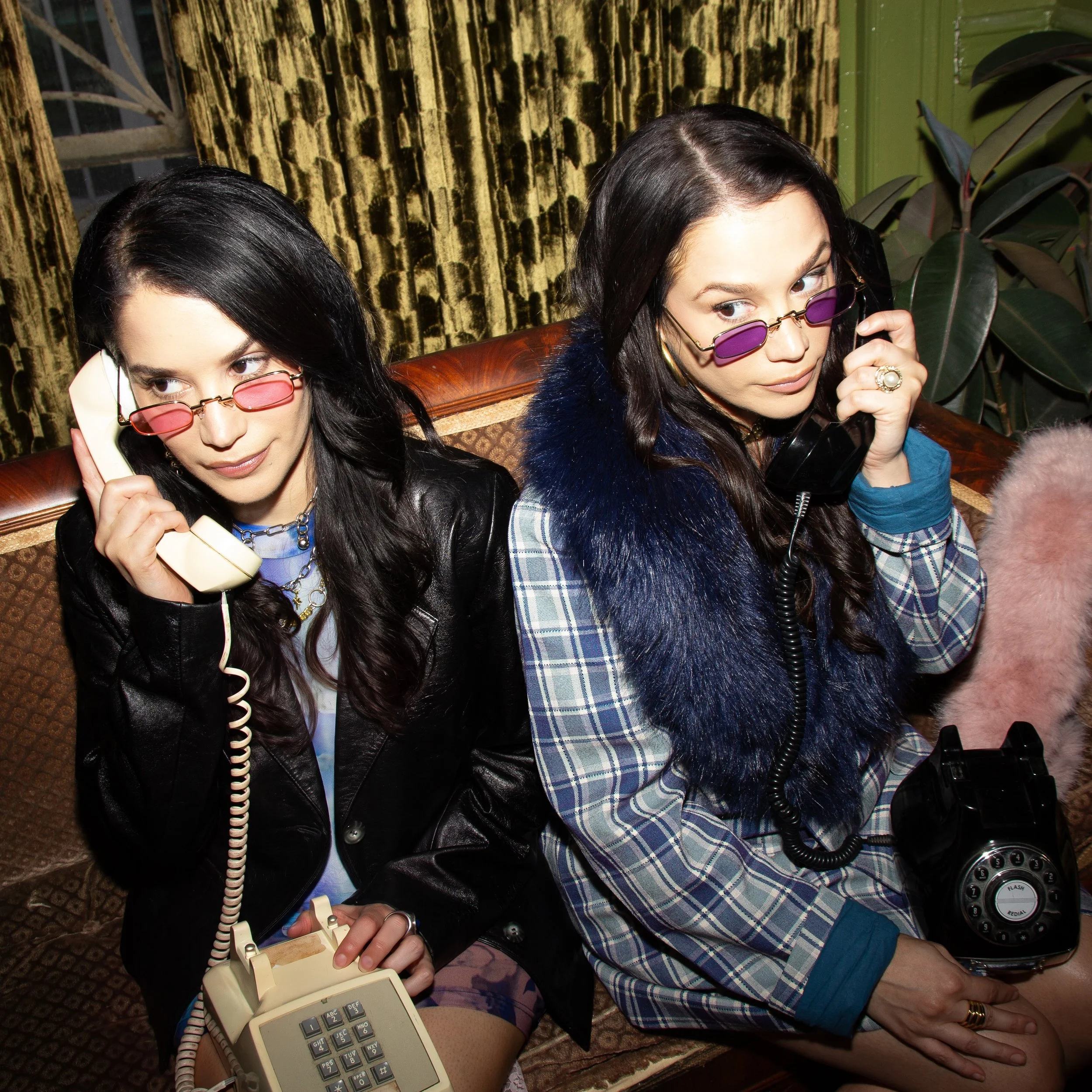 Two women with dark hair and purple tinted sunglasses talking on vintage landline phones in a retro-themed room with patterned velvet curtains and green walls, sitting close together at a wooden table.