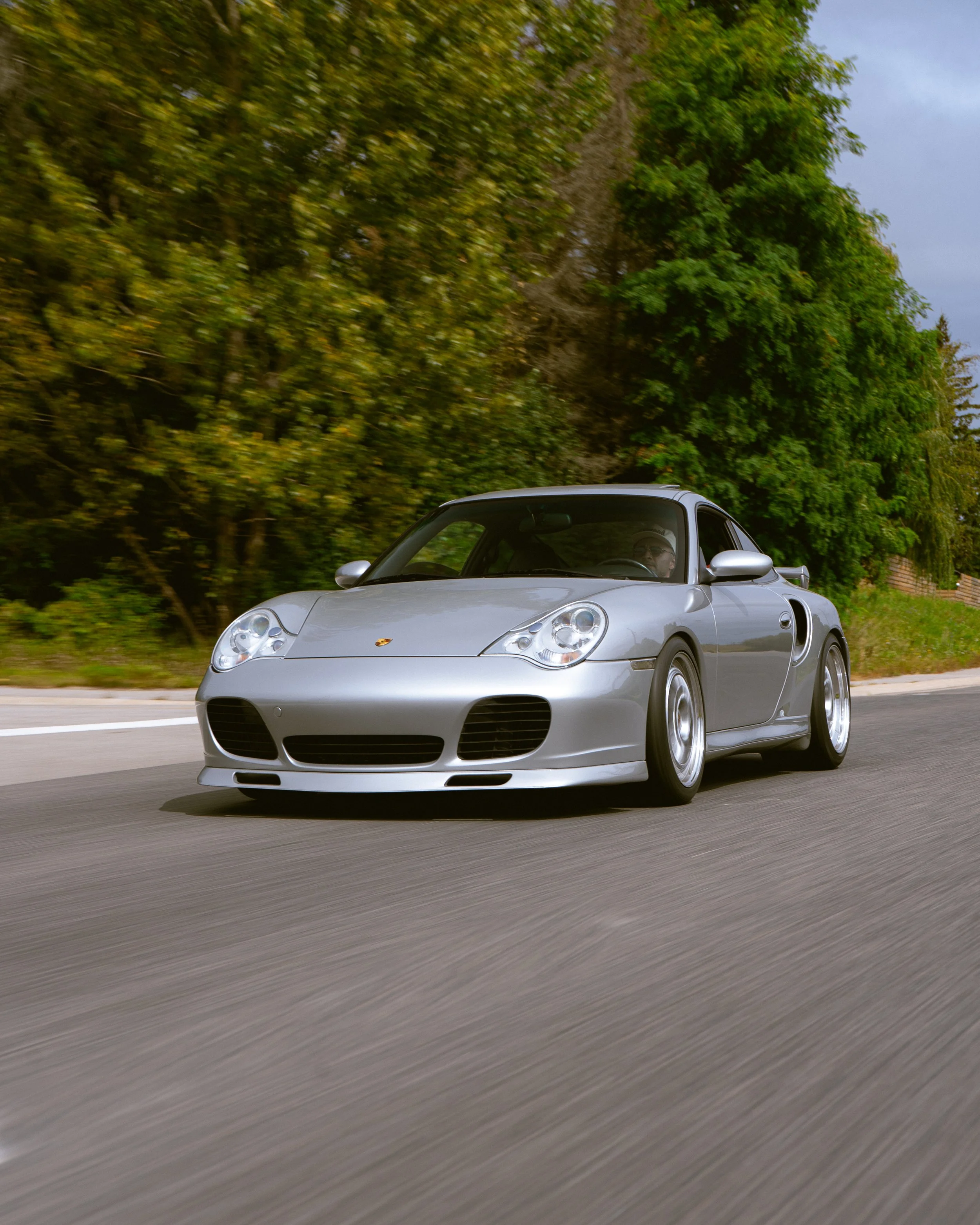 A silver Porsche sports car driving on a paved road with trees and greenery in the background.