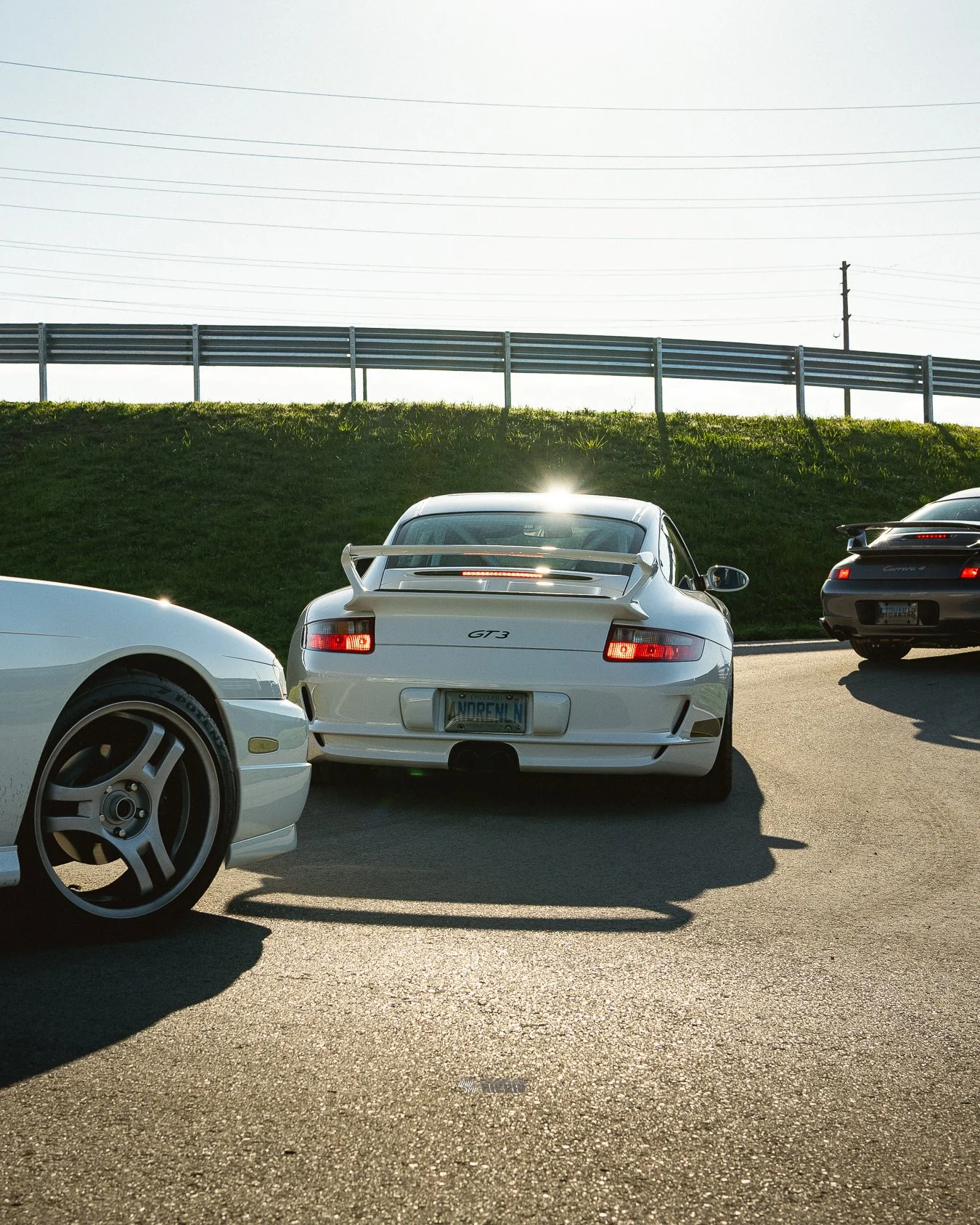 Three cars parked near a grassy hill with a guardrail and power lines in the background. The car in the middle is a white Porsche 911 GT3 with a spoiler, and the other two cars are white and black sports cars.