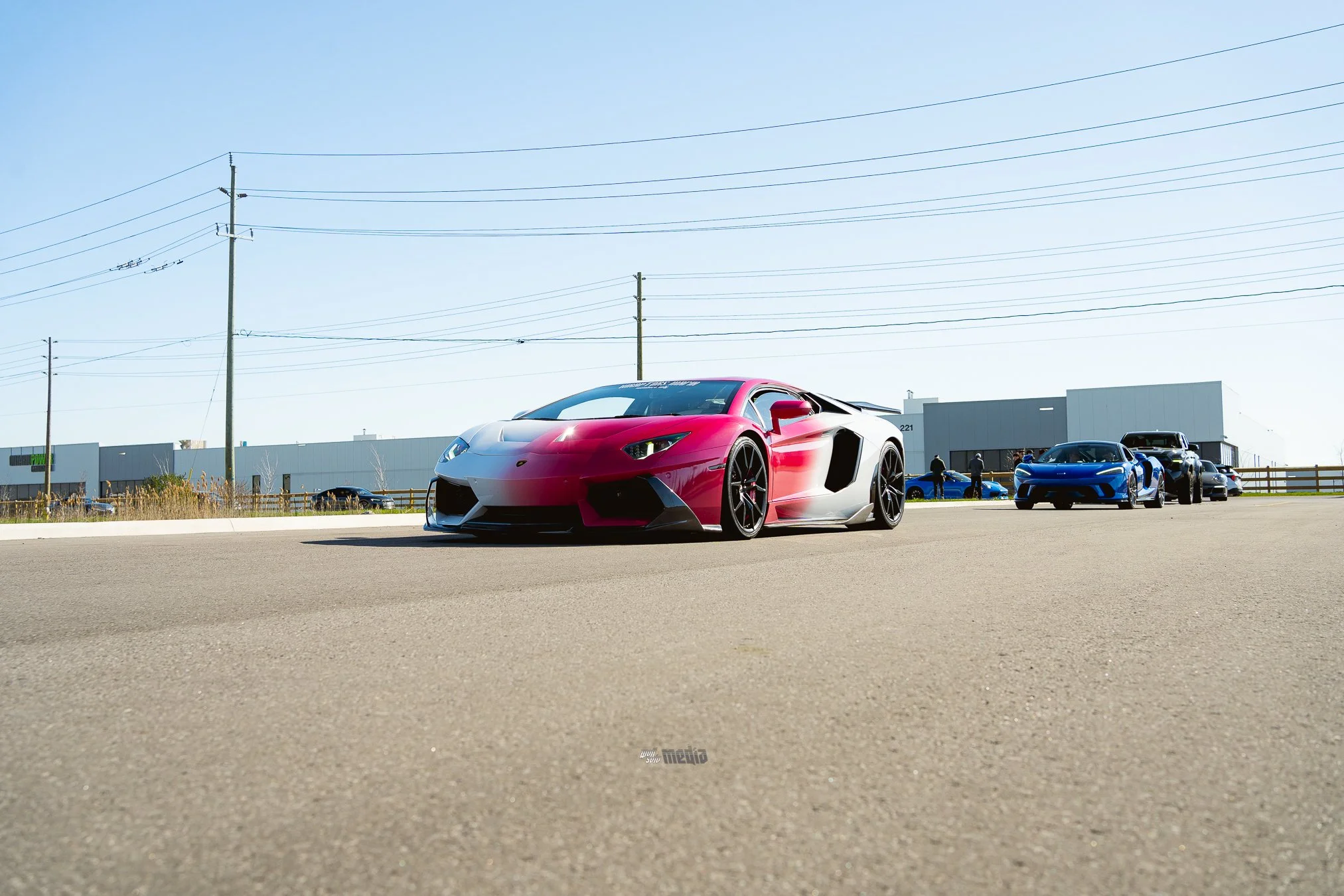 A pink and white sports car parked in a lot with other cars and a building in the background, under a clear blue sky.