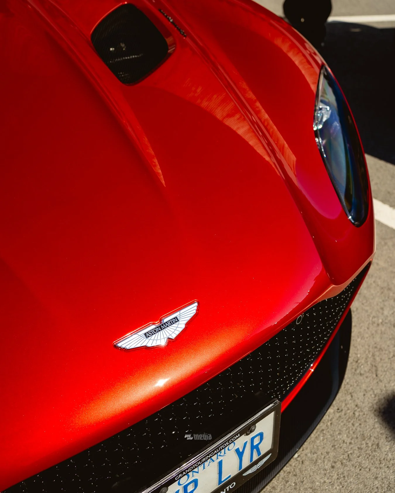 Close-up of a red Aston Martin sports car's front end, showing the vehicle's badge, headlight, and front grille, with a partial view of the license plate.