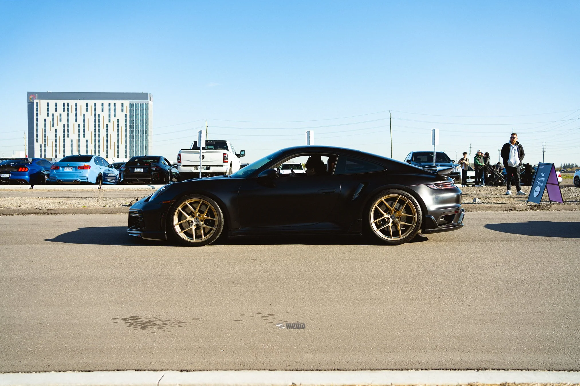 Black sports car with gold rims parked in a parking lot, with other cars and people in the background under a clear blue sky.