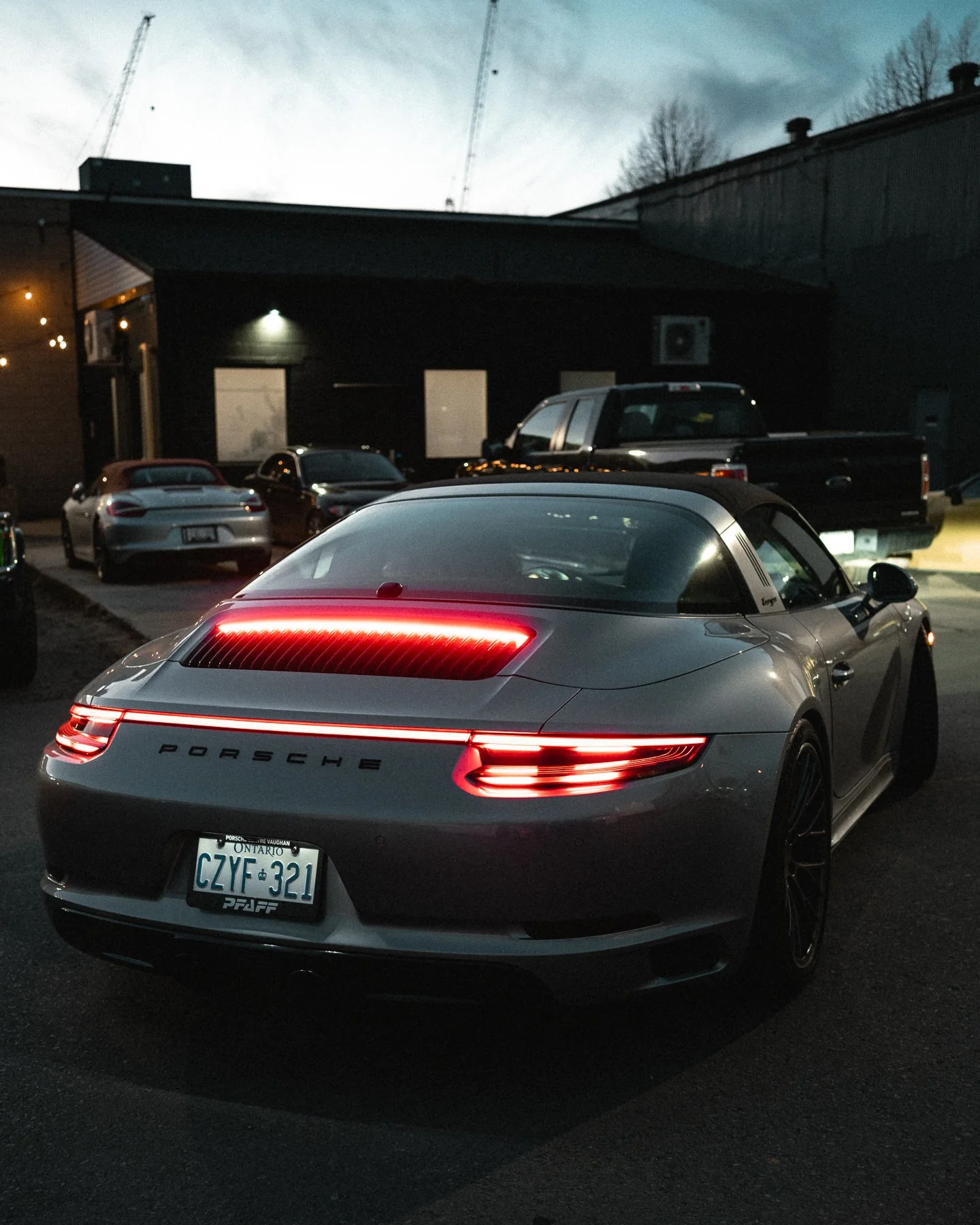 A silver Porsche convertible parked in a dark parking lot during evening or night, with multiple other vehicles and a building in the background.