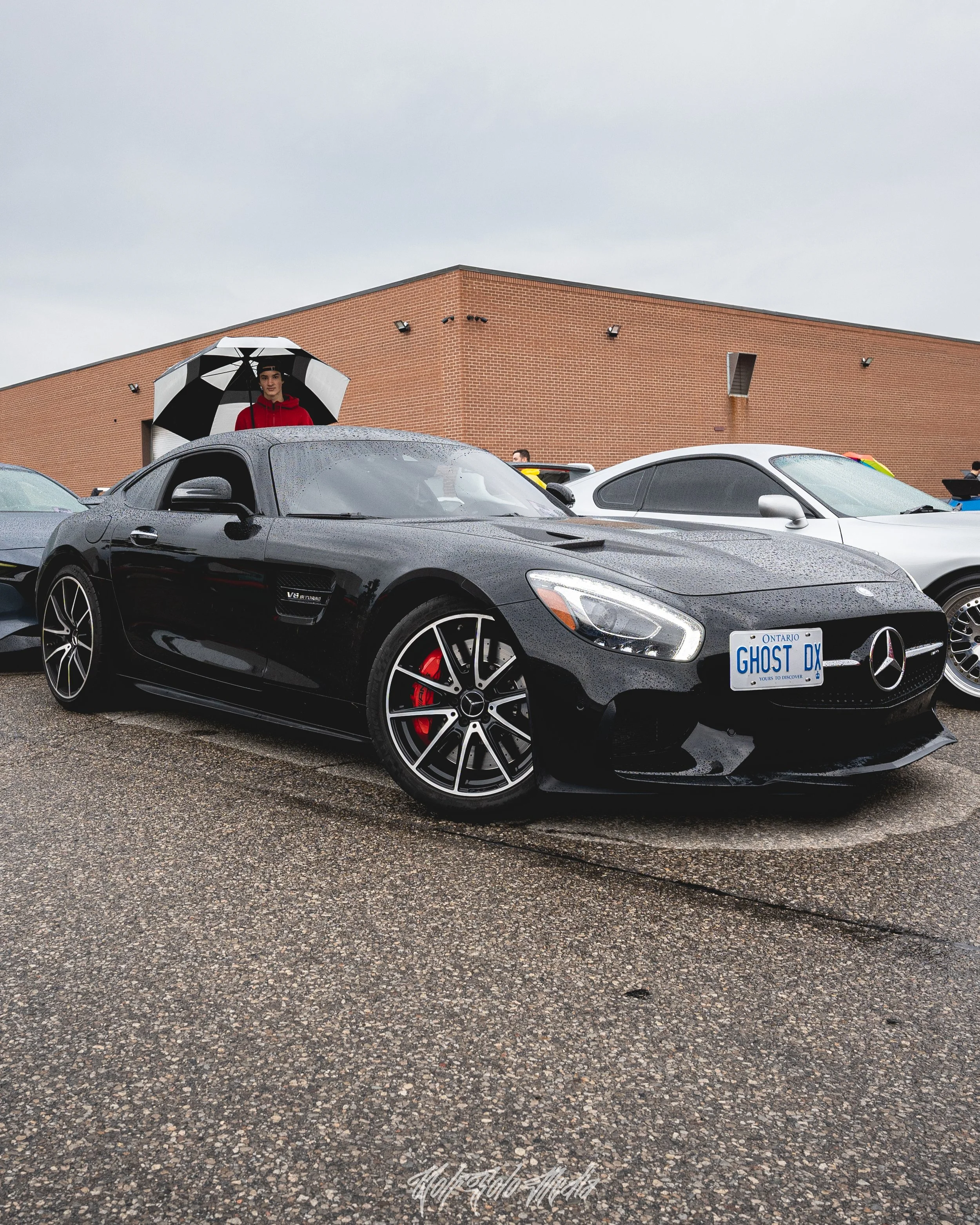 A black Mercedes-Benz SLS AMG sports car parked in a lot with other vehicles on a rainy day, with a person holding a black and white umbrella standing behind it.