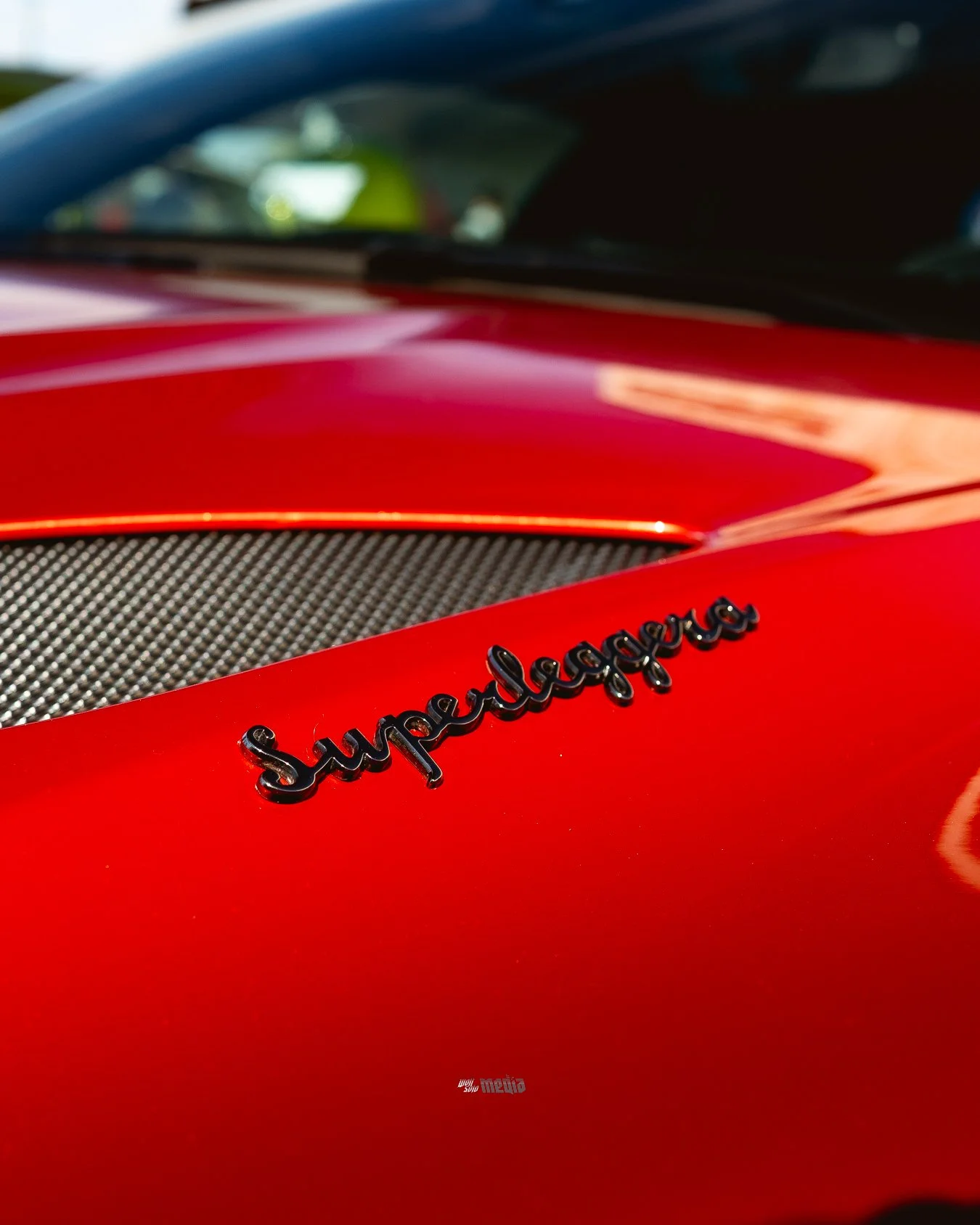 Close-up of a red sports car with a black 'Superleggera' emblem on the hood, showing part of the windshield and black carbon fiber component.