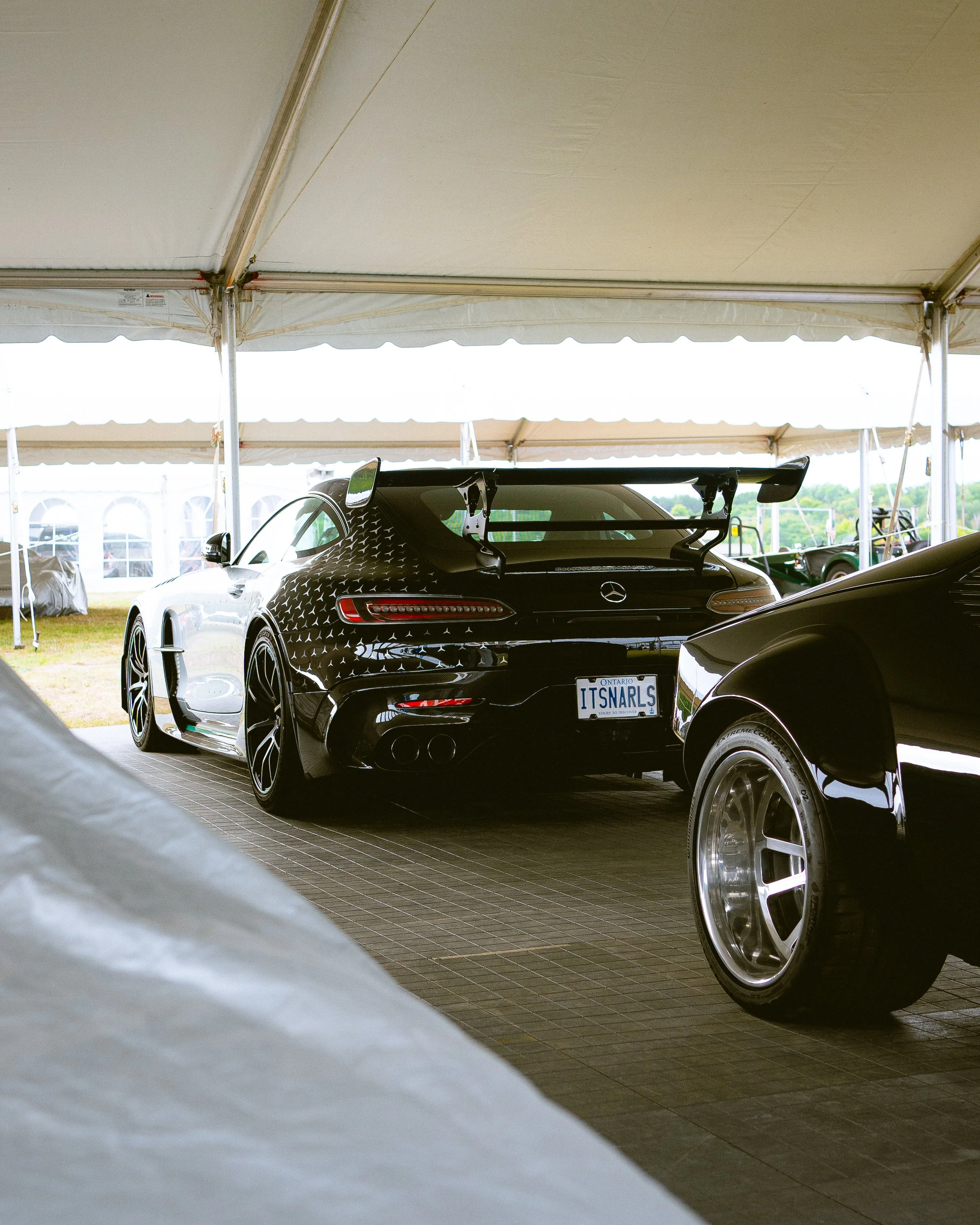 Rear view of a black Mercedes-Benz sports car with a large spoiler, parked inside a tent at what appears to be a car show or event.