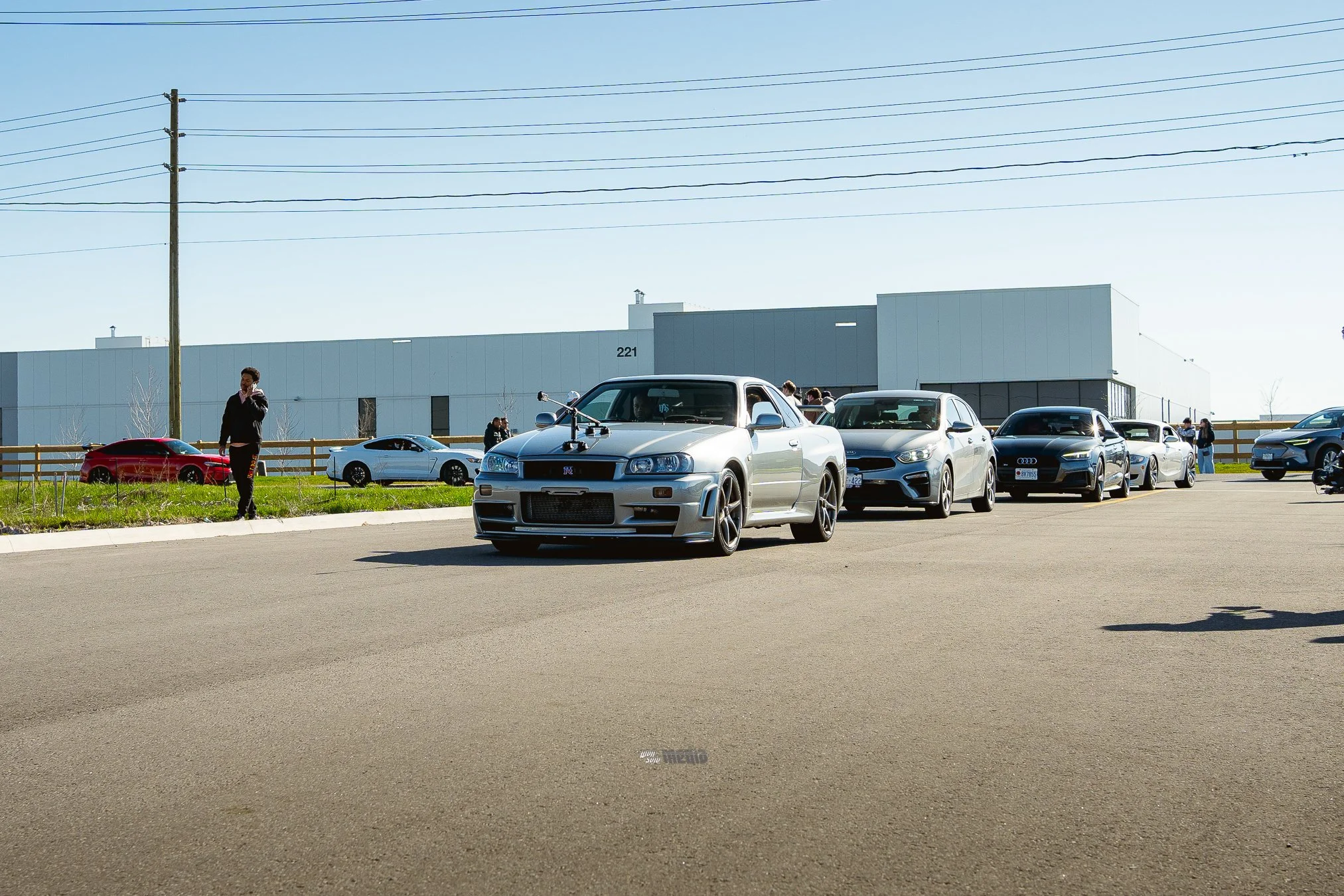 View of a parking lot with several parked cars, including a silver Nissan Skyline R34 in the foreground. There are people walking and talking, with a modern industrial building with the number 221 in the background. The sky is clear and blue.