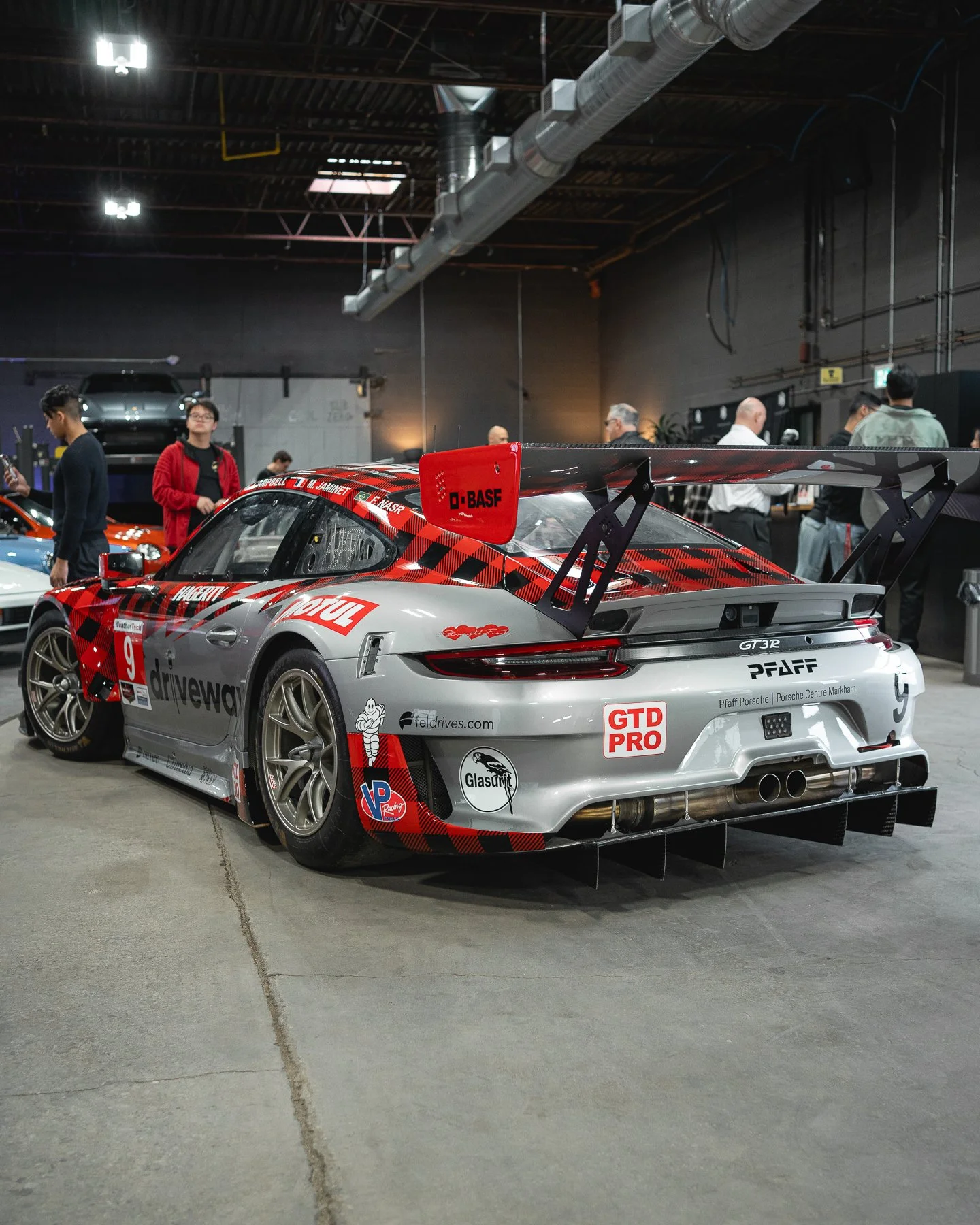 Grey racing Porsche 911 GT3 R with red and black checkered accents, large rear wing, and sponsorship stickers displayed indoors at a car event.