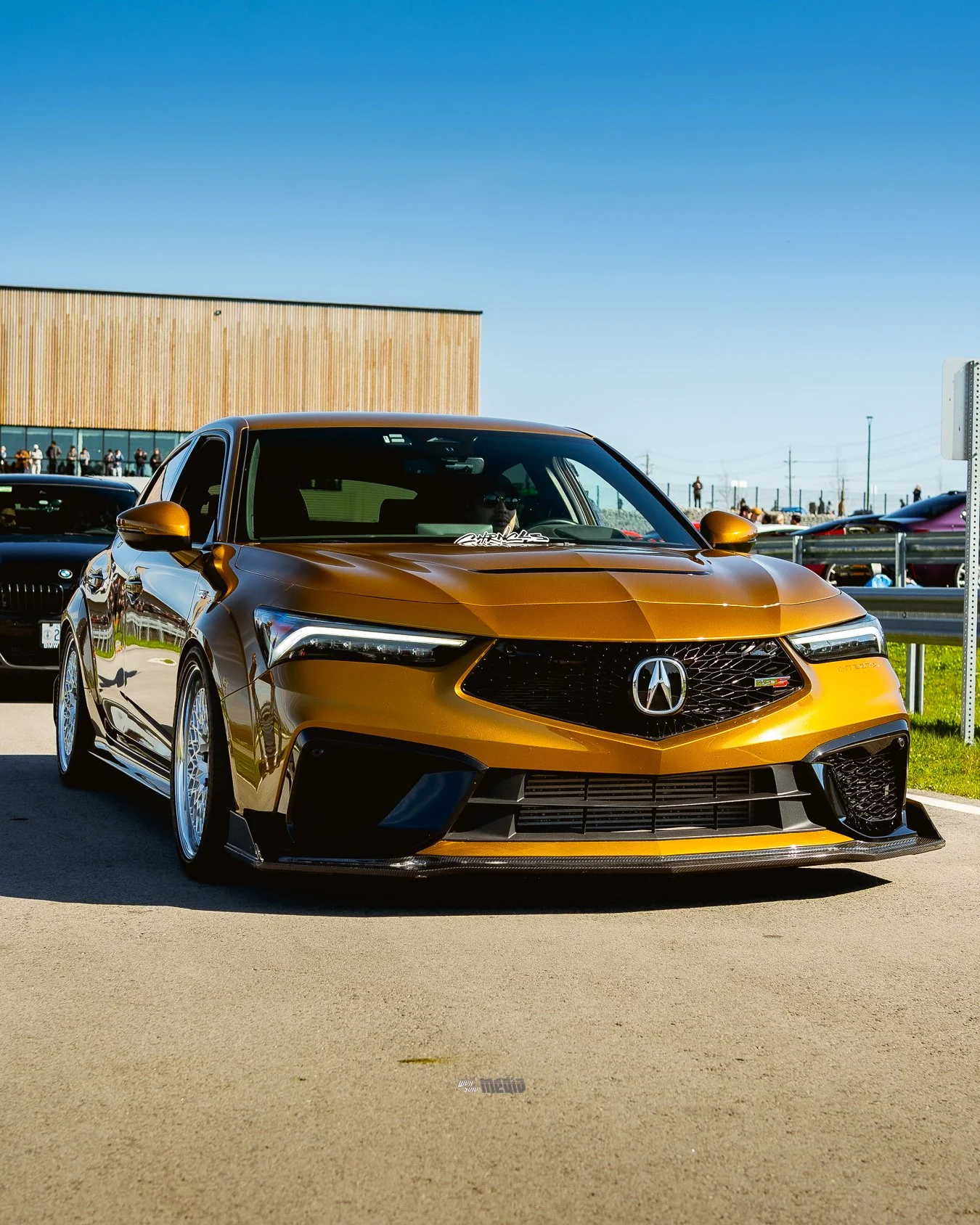 A metallic gold Acura sports car parked outdoors on a sunny day, with other cars and a building with vertical wooden panels in the background.