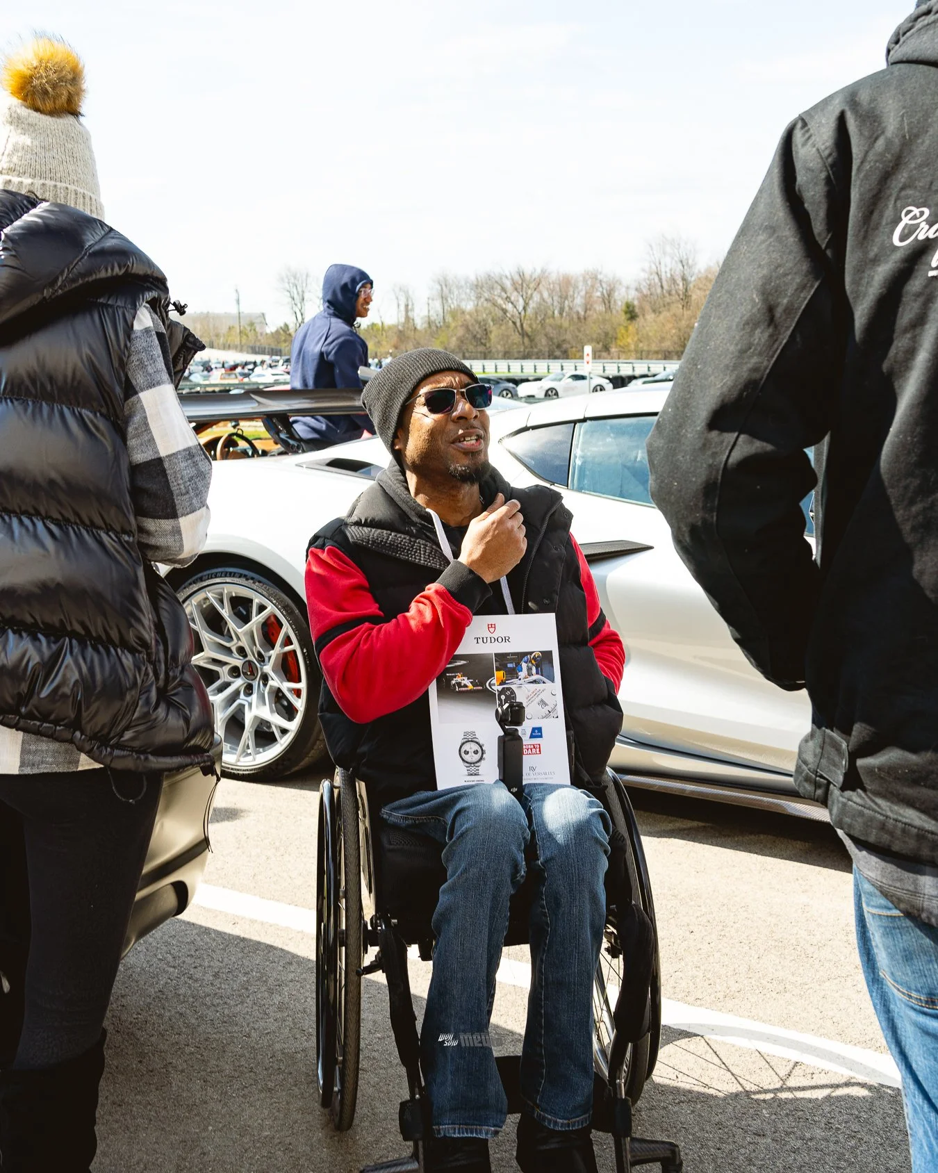 A man in a wheelchair wearing sunglasses and a beanie engaging in conversation with others near a white sports car, with additional people and cars visible in the background outdoors on a sunny day.