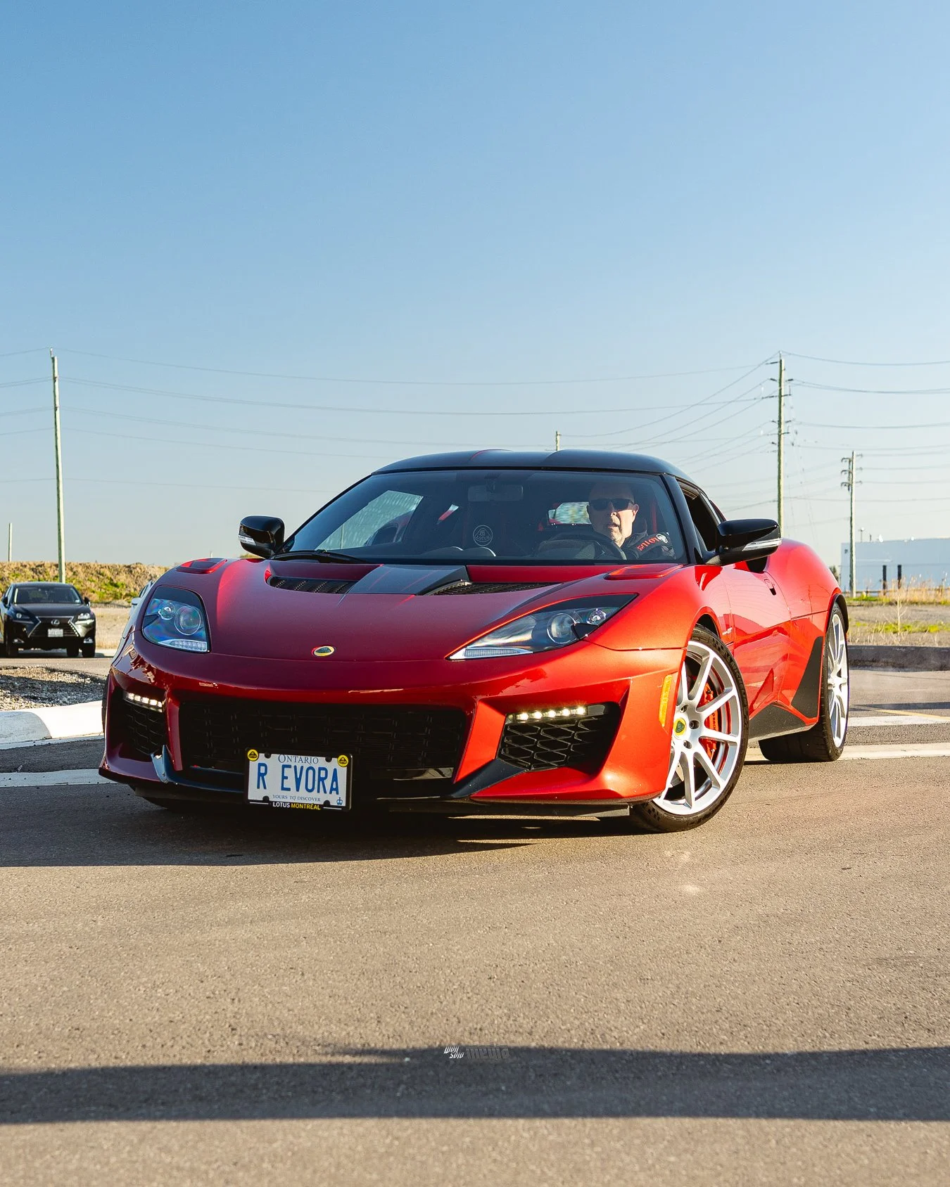 A red sports car with a black hood, parked outdoors under clear blue skies. The car has a custom Ontario license plate reading "R EVORA" and features white alloy wheels. A person wearing sunglasses is sitting inside the car.
