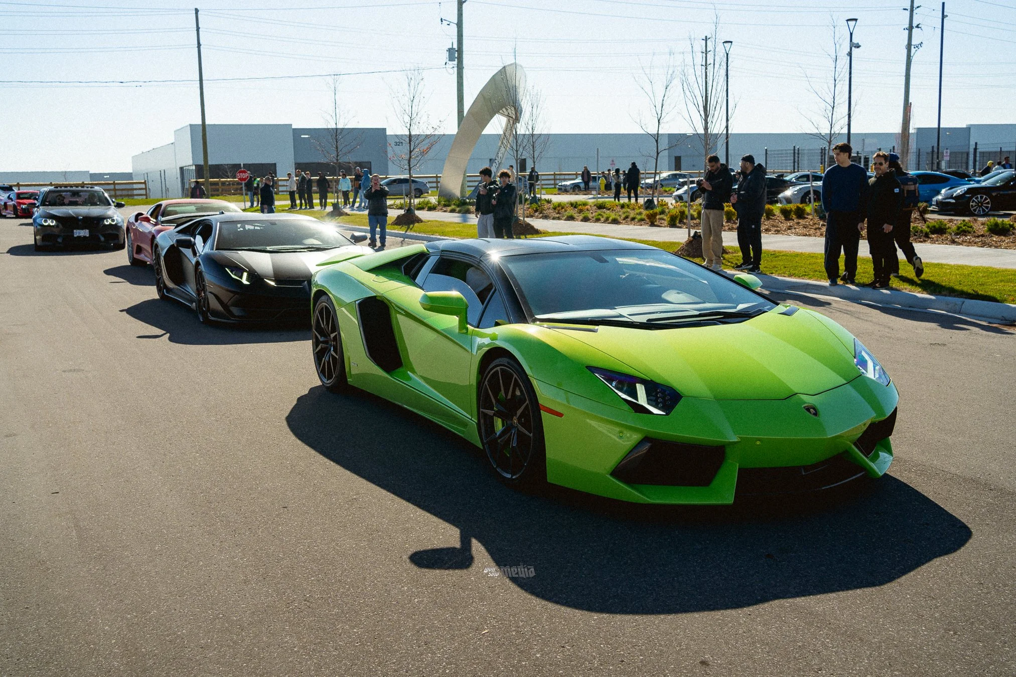 Several luxury sports cars, including a green Lamborghini and a black Lamborghini, parked outdoors with a small crowd of people gathered around and taking photos. There are a few spectators near a sculpture and a parking lot in the background.