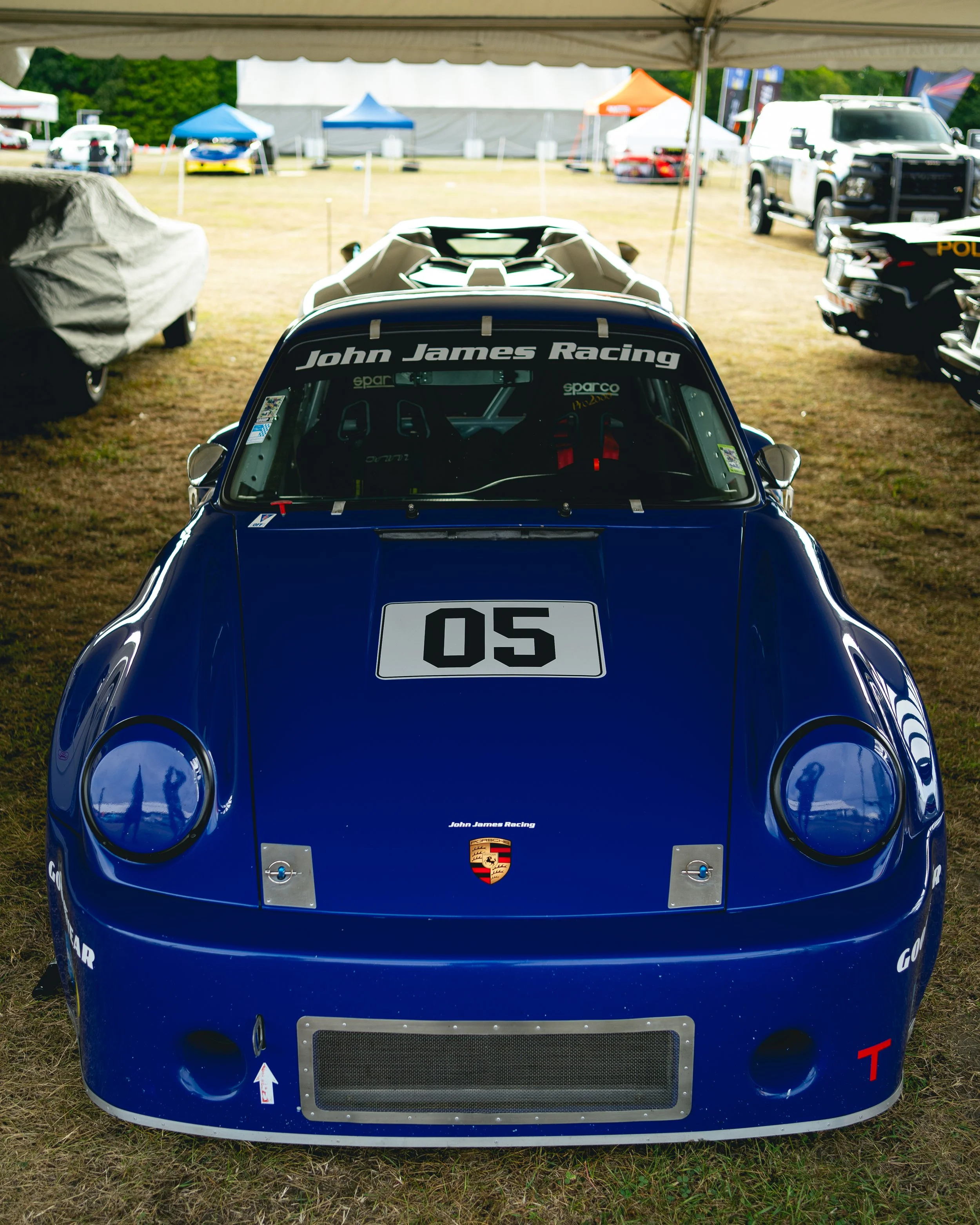 Blue racing car with number 05 and Porsche logo, parked under a tent at a race event.