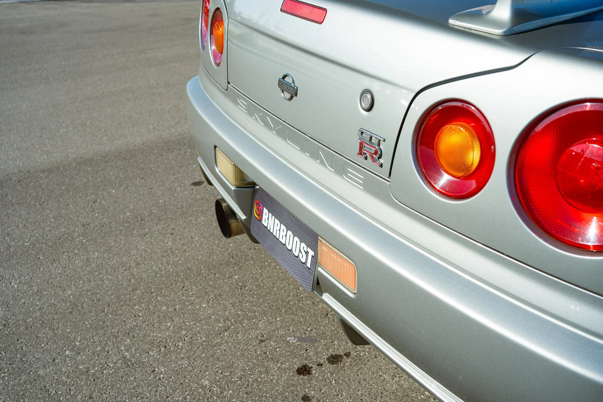 Close-up of the rear of a silver Nissan Skyline GTR car, showcasing round taillights, badges, and exhaust pipe on an asphalt surface.