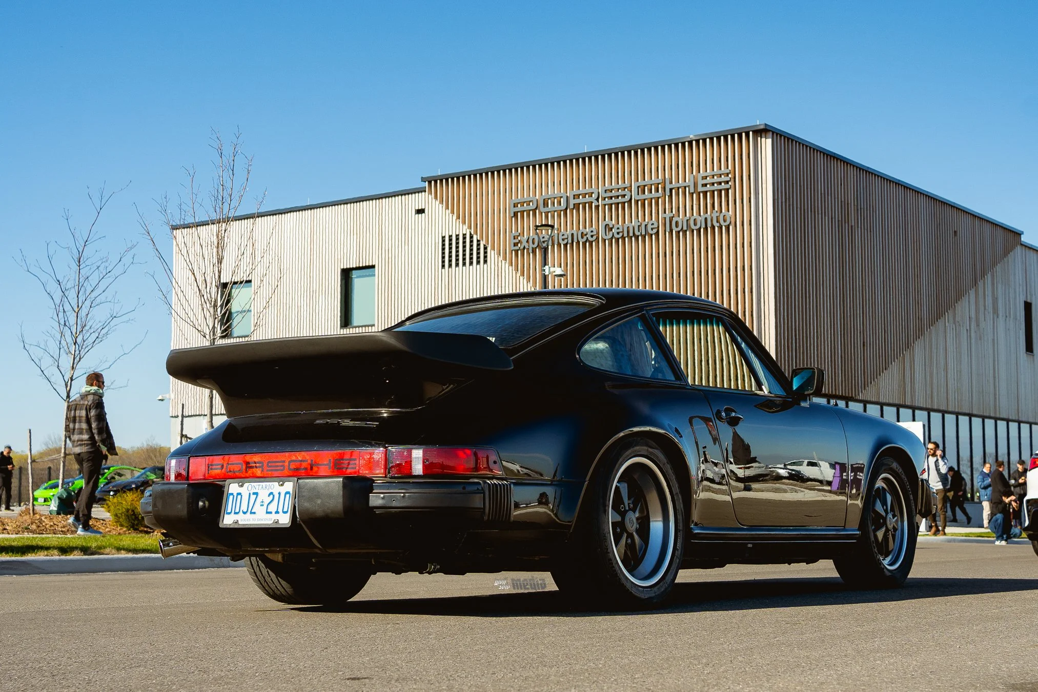 Black Porsche sports car parked outdoors near a modern building with wooden siding. People are walking in the background on a clear day with a blue sky.