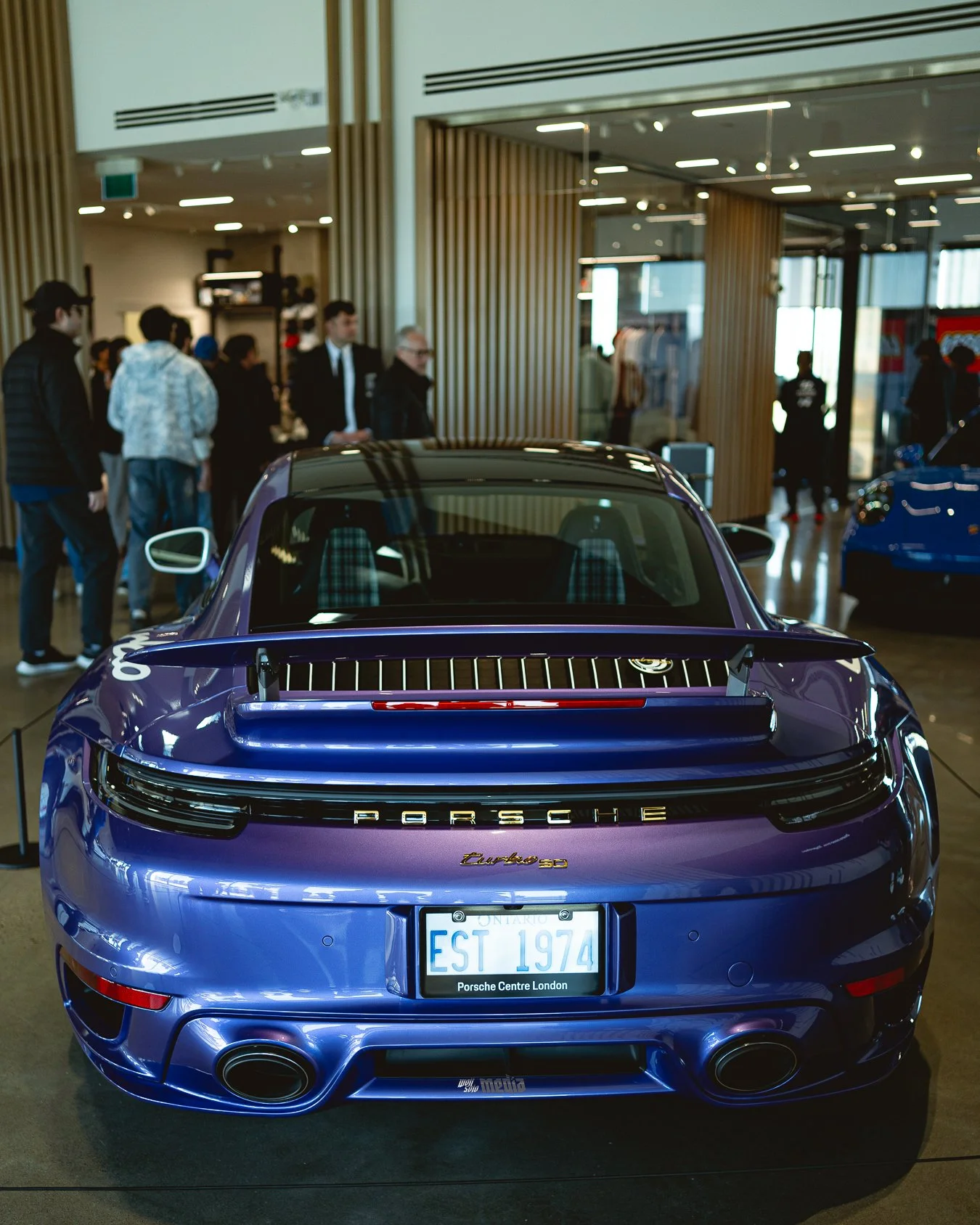 A purple Porsche car on display at an indoor auto show, with people observing in the background.