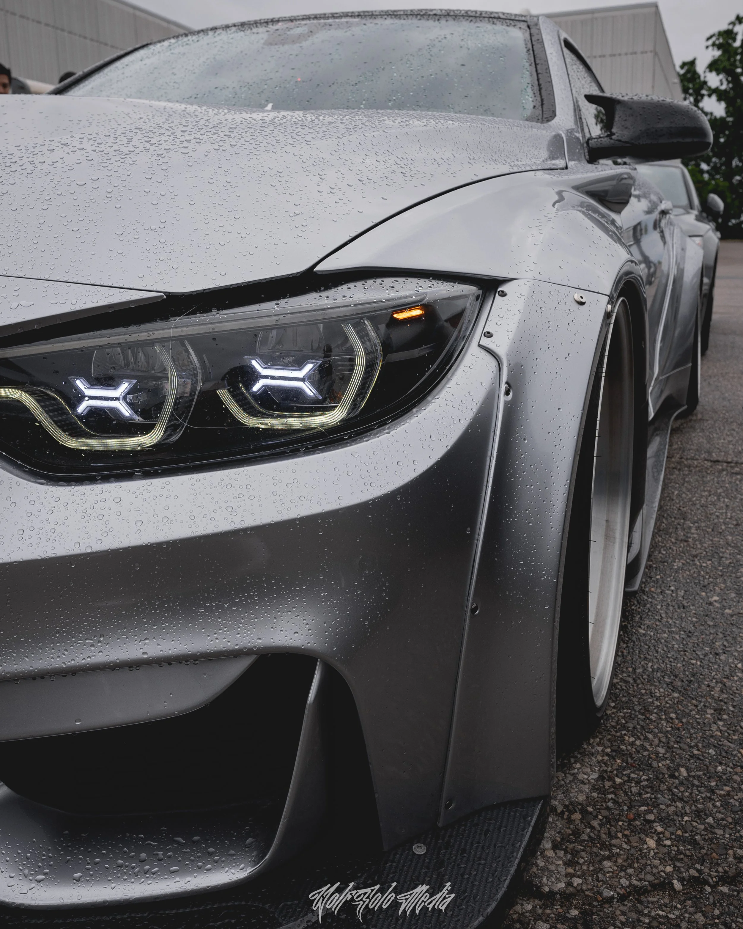 Close-up of a silver sports car with rain droplets on its surface, featuring distinctive LED headlights and a black front splitter.