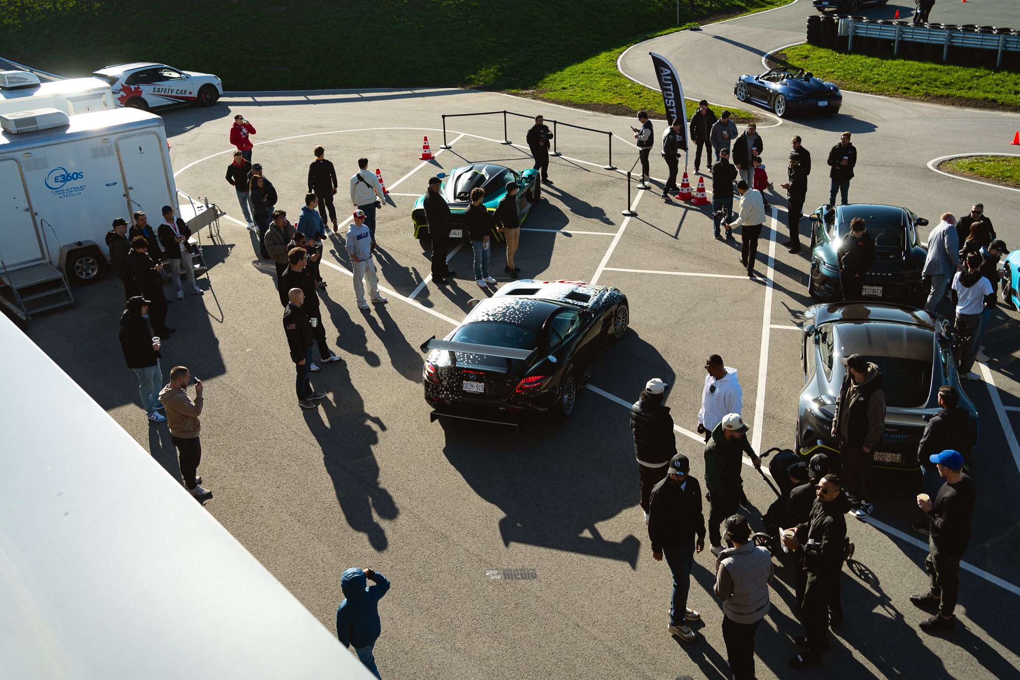 A group of people gathered around sports cars at a car event, with some cars parked on the pavement and others on a racetrack. There are banners, cones, and a trailer, with people socializing and taking pictures.