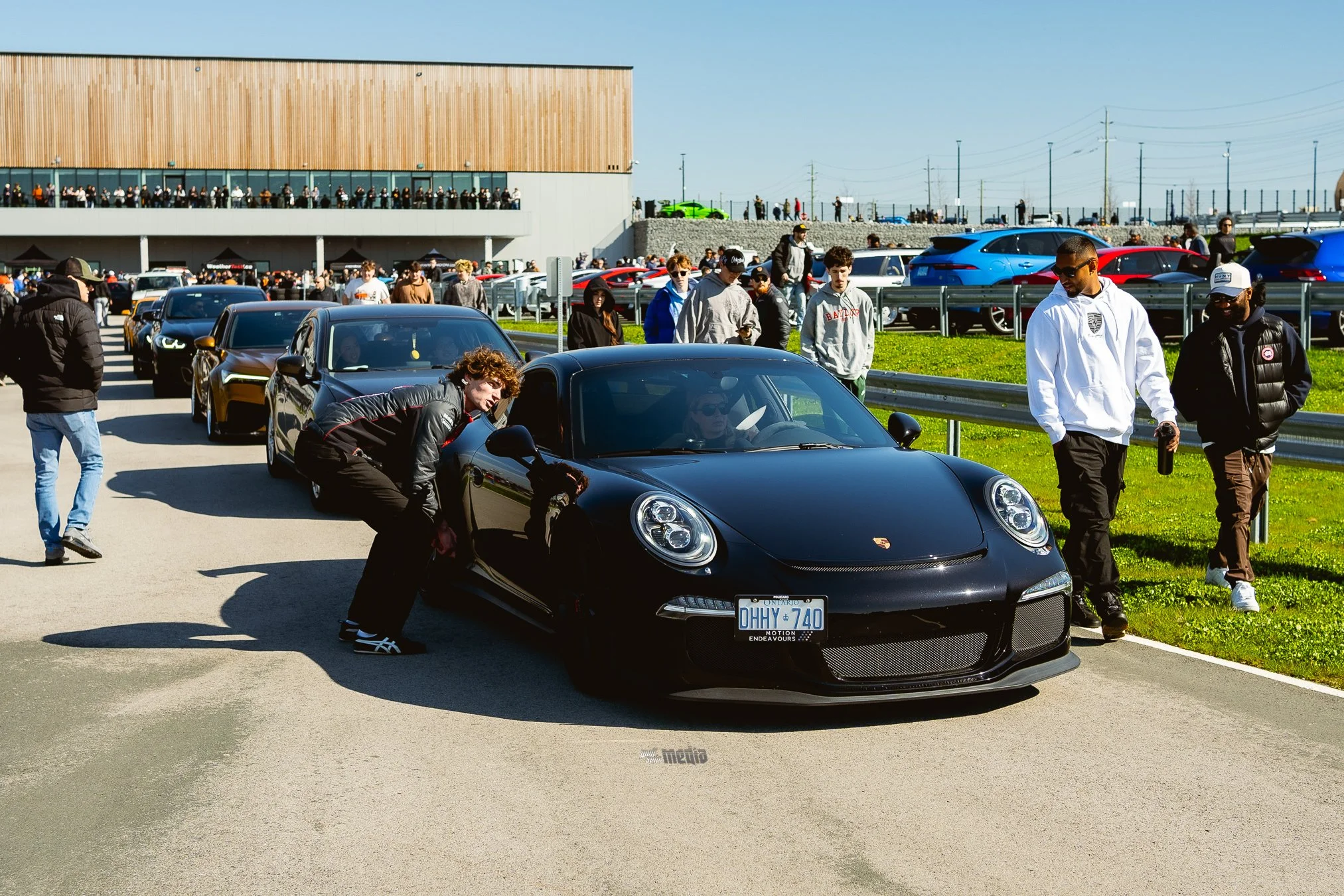 A lineup of cars, including a black Porsche, with a crowd of people gathered around them at an outdoor car event on a sunny day. In the background, there is a large building with a balcony full of spectators and more cars parked on a hill.