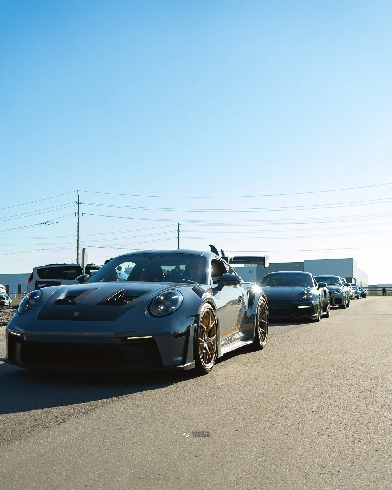 Line of high-performance sports cars, including a silver Porsche 911 GT2RS, parked in a lot under a clear blue sky, with industrial buildings and power lines in the background.