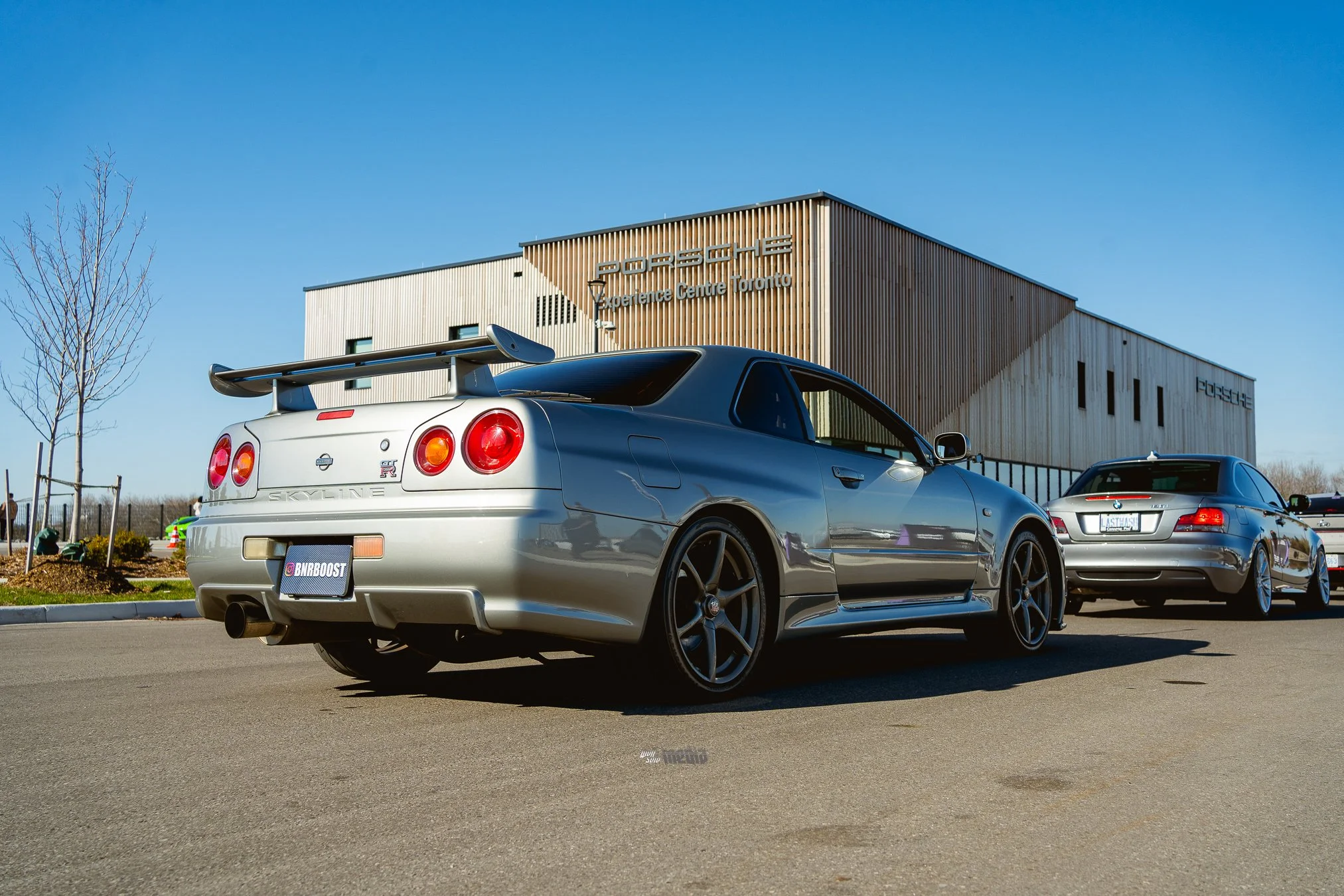 A silver Nissan Skyline GT-R parked in a lot near a Porsche Experience Centre Toronto building with a wooden exterior. The sky is clear and blue, with a small leafless tree and other cars visible in the background.