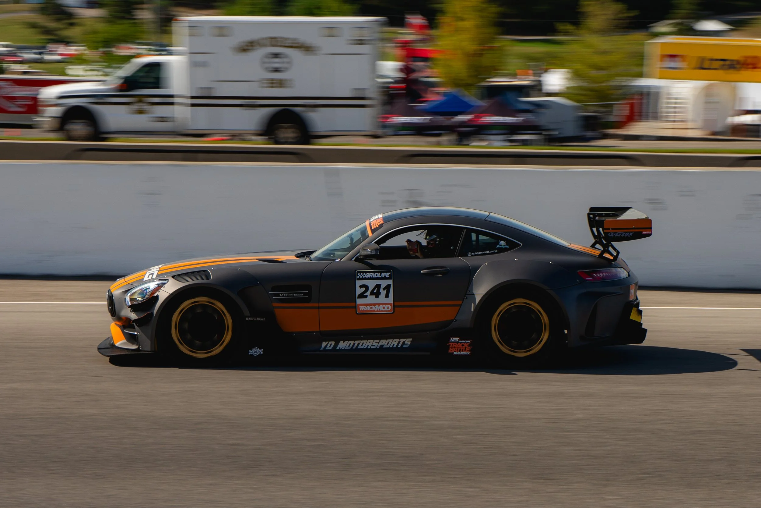 A black racing car with orange accents and the number 241 on the side speeds along a race track, with a blurred background of trailers and tents.