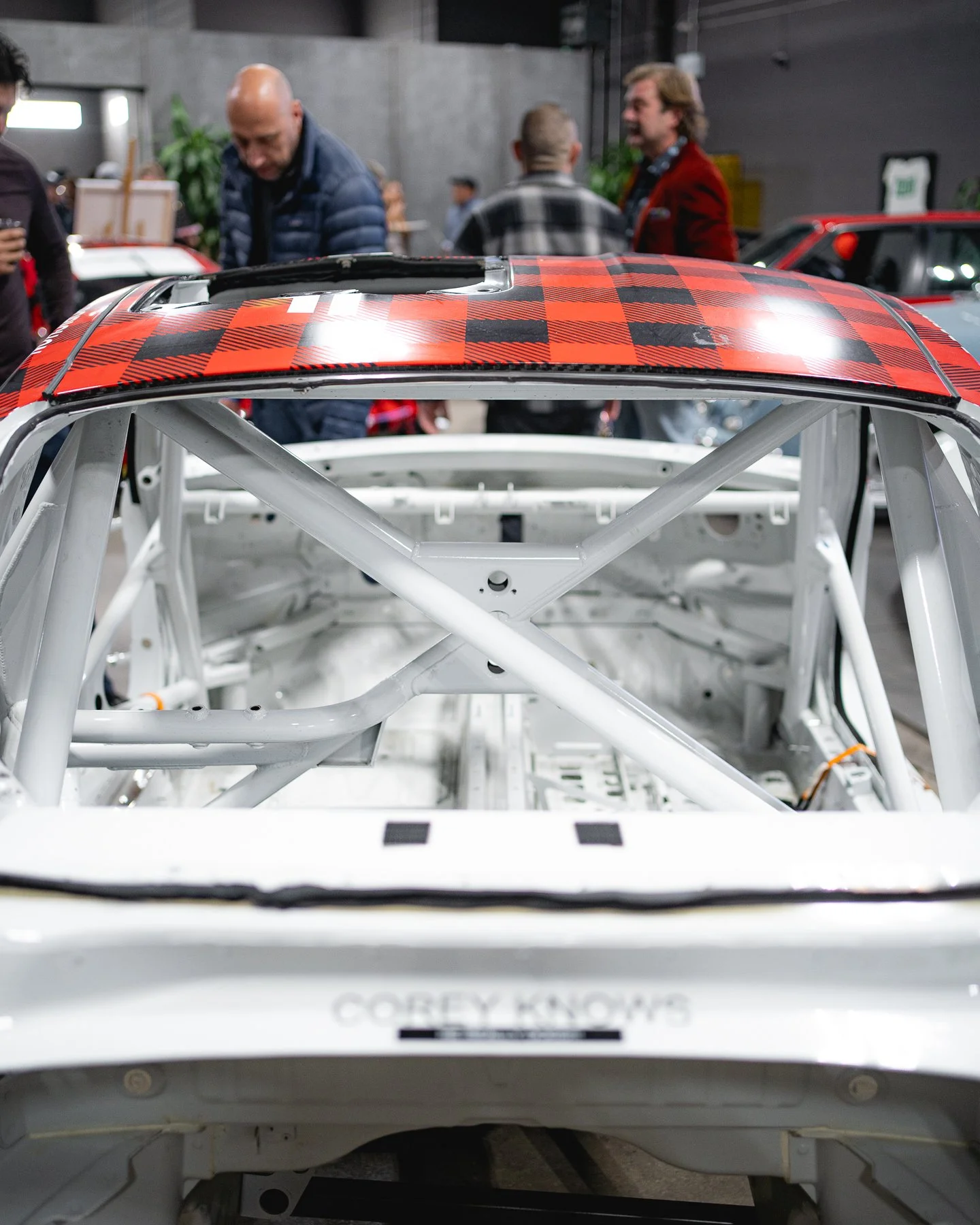 Inside view of a race car with a red and black checkered roof, showing the car's white roll cage and chassis, with people observing in the background at an indoor car event.