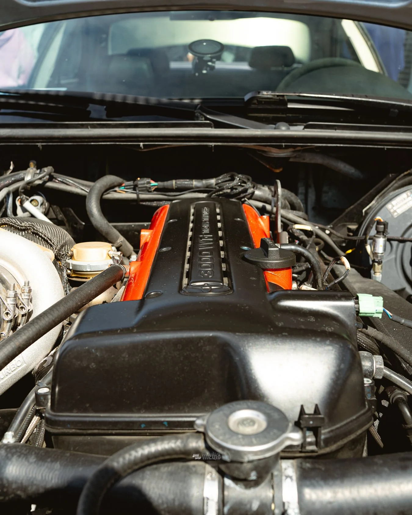 Car engine with visible black and orange engine cover and various hoses and components, viewed from the front of the engine bay.