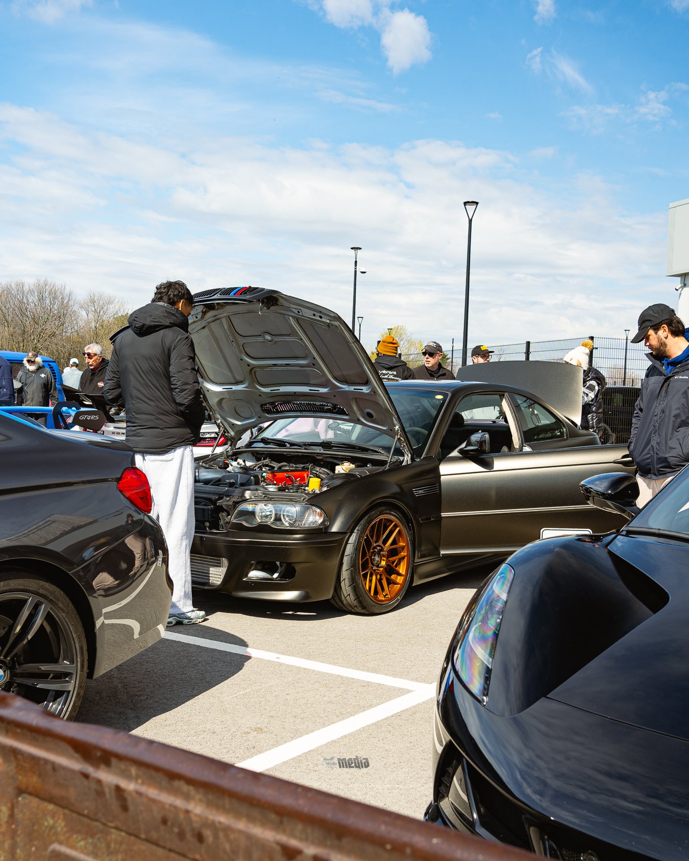 Group of people at a car meet inspecting a black sports car with its hood open, parked in a lot on a partly cloudy day.