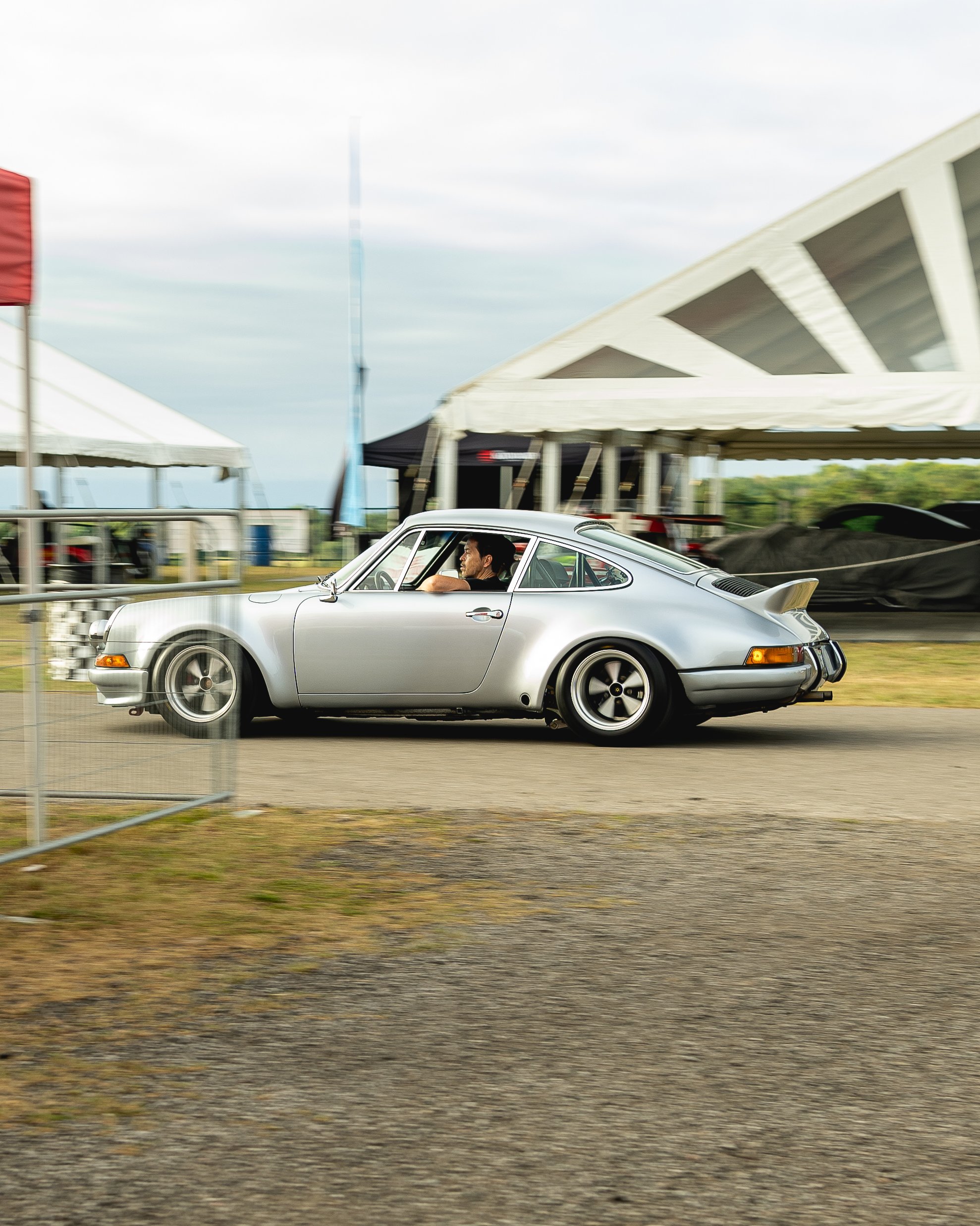 Silver vintage Porsche 911 sports car driving past tents at an outdoor event.