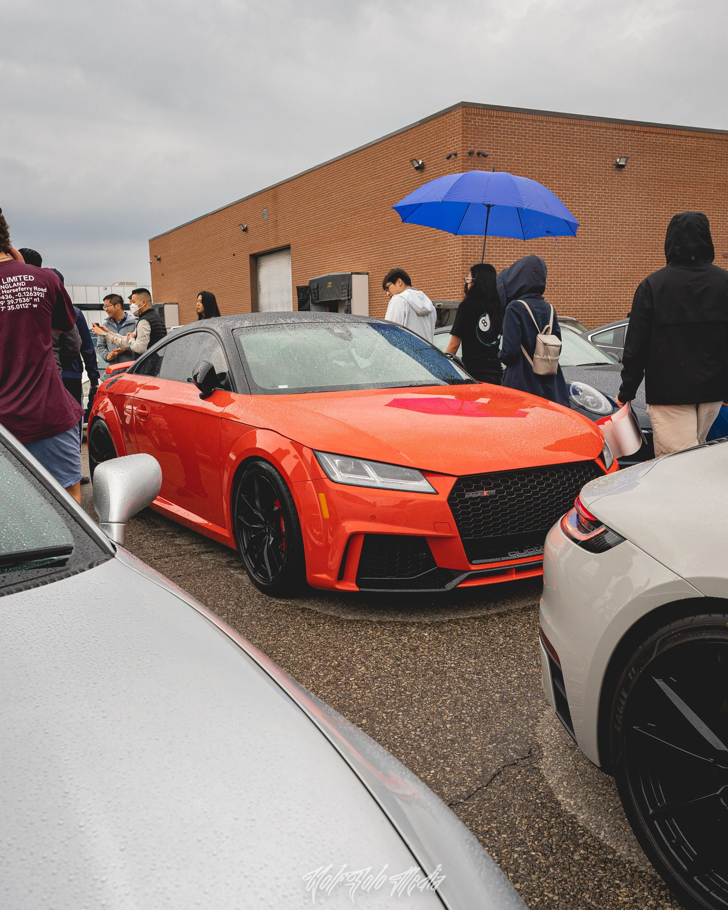 Red sports car parked in a lot on a rainy day, surrounded by people, some holding umbrellas, near a brick building.
