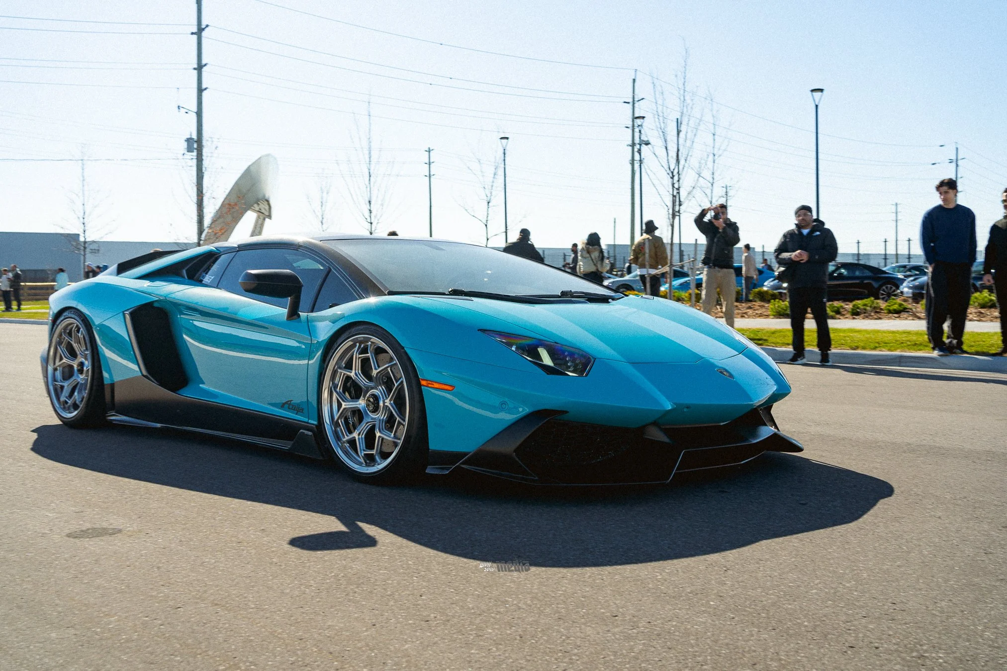 A blue Lamborghini sports car parked at an outdoor car show with people walking around and taking photos in the background.