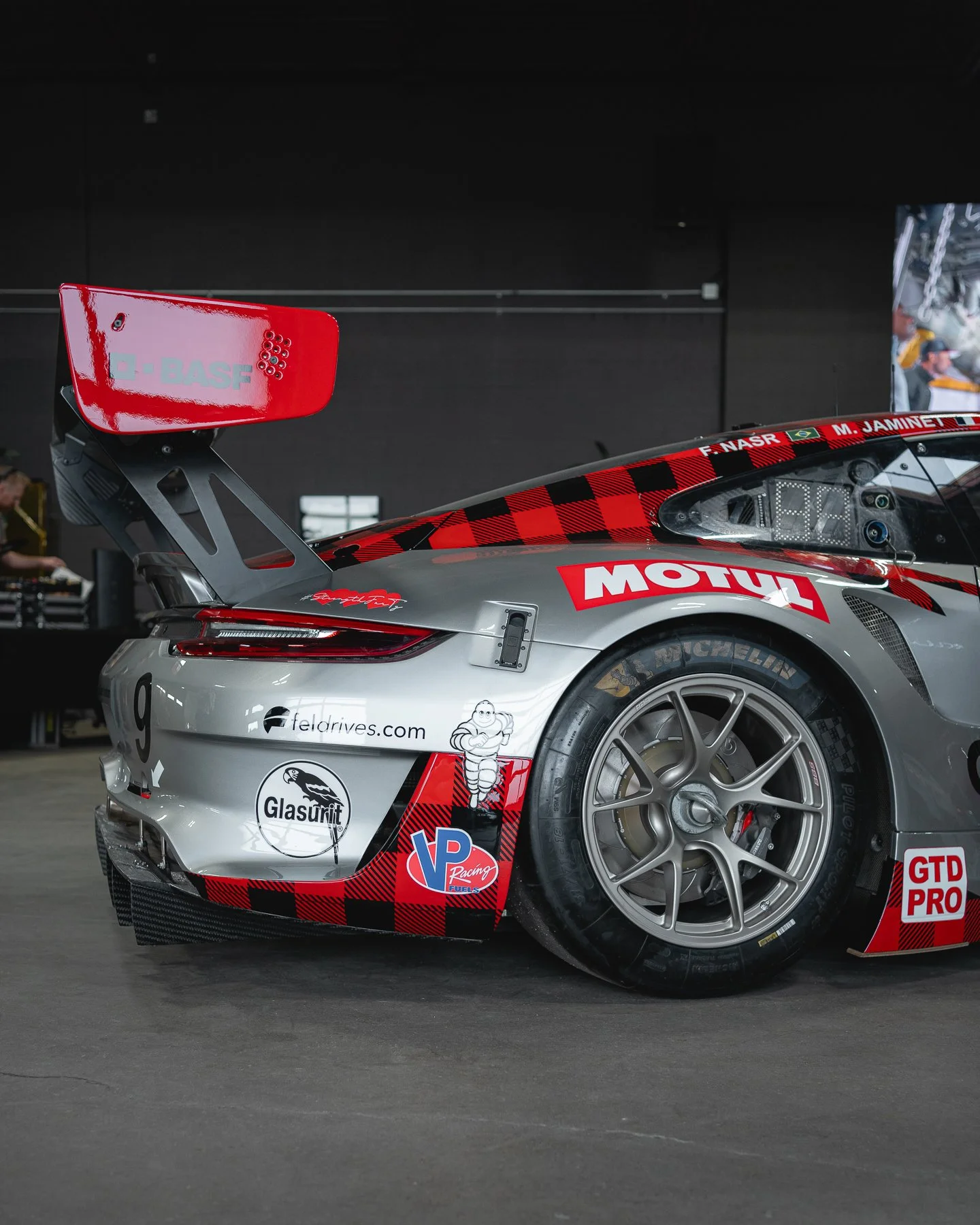Close-up of a race car's rear section, featuring a large rear wing, racing tires, sponsorship logos, and a sleek aerodynamic design.