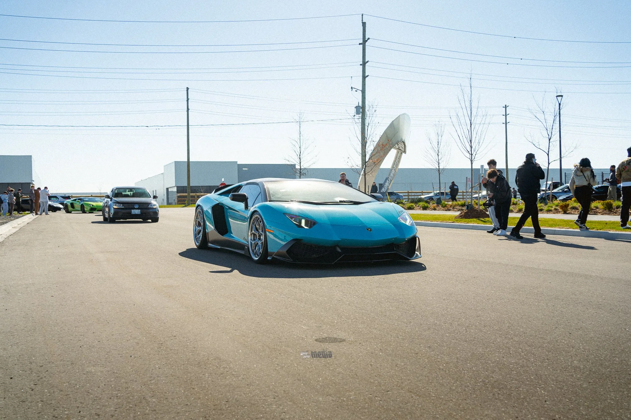A blue luxury sports car parked in an outdoor area with several people and other cars in the background.