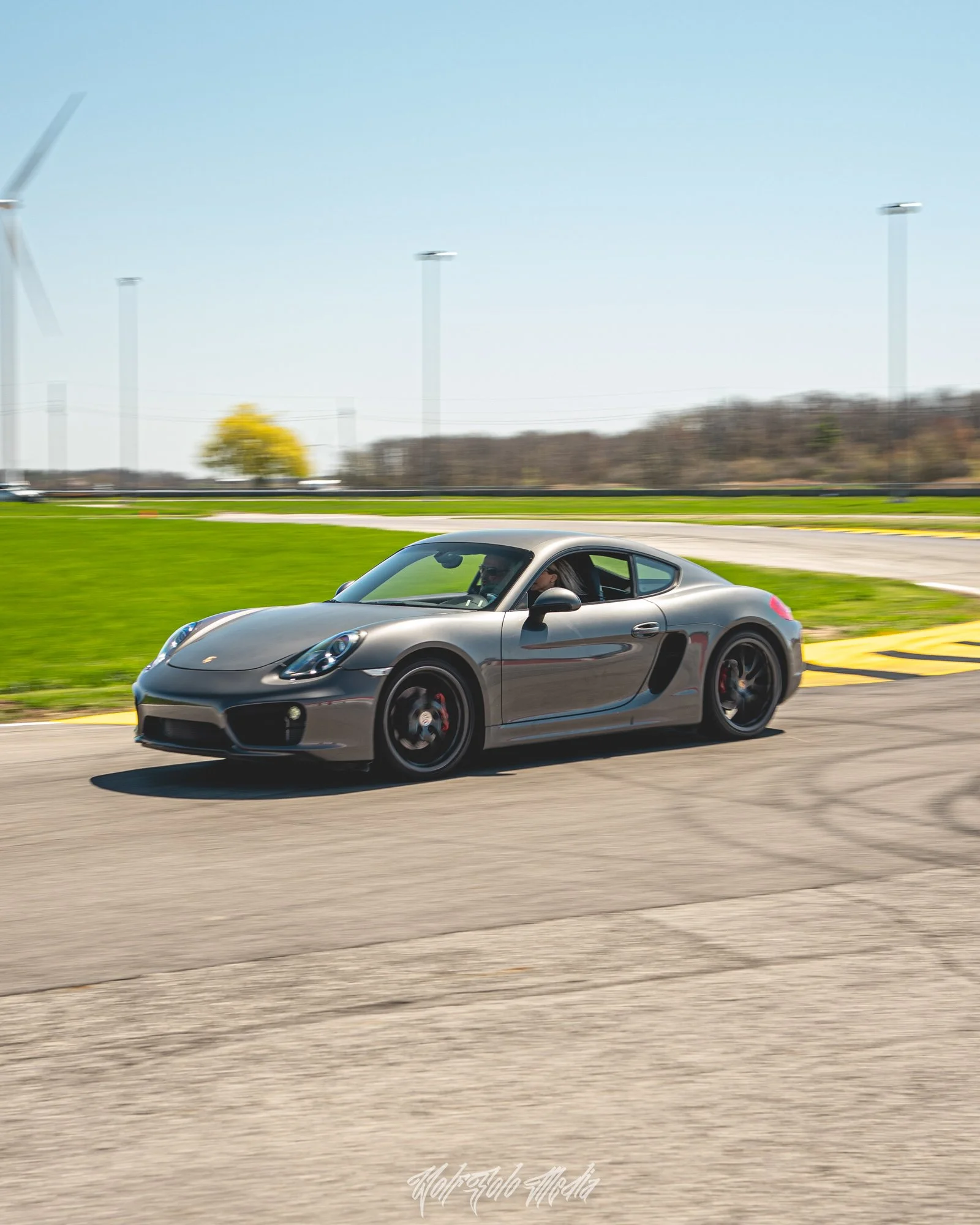 A silver Porsche sports car driving on a race track with green grass and wind turbines in the background under a clear blue sky.