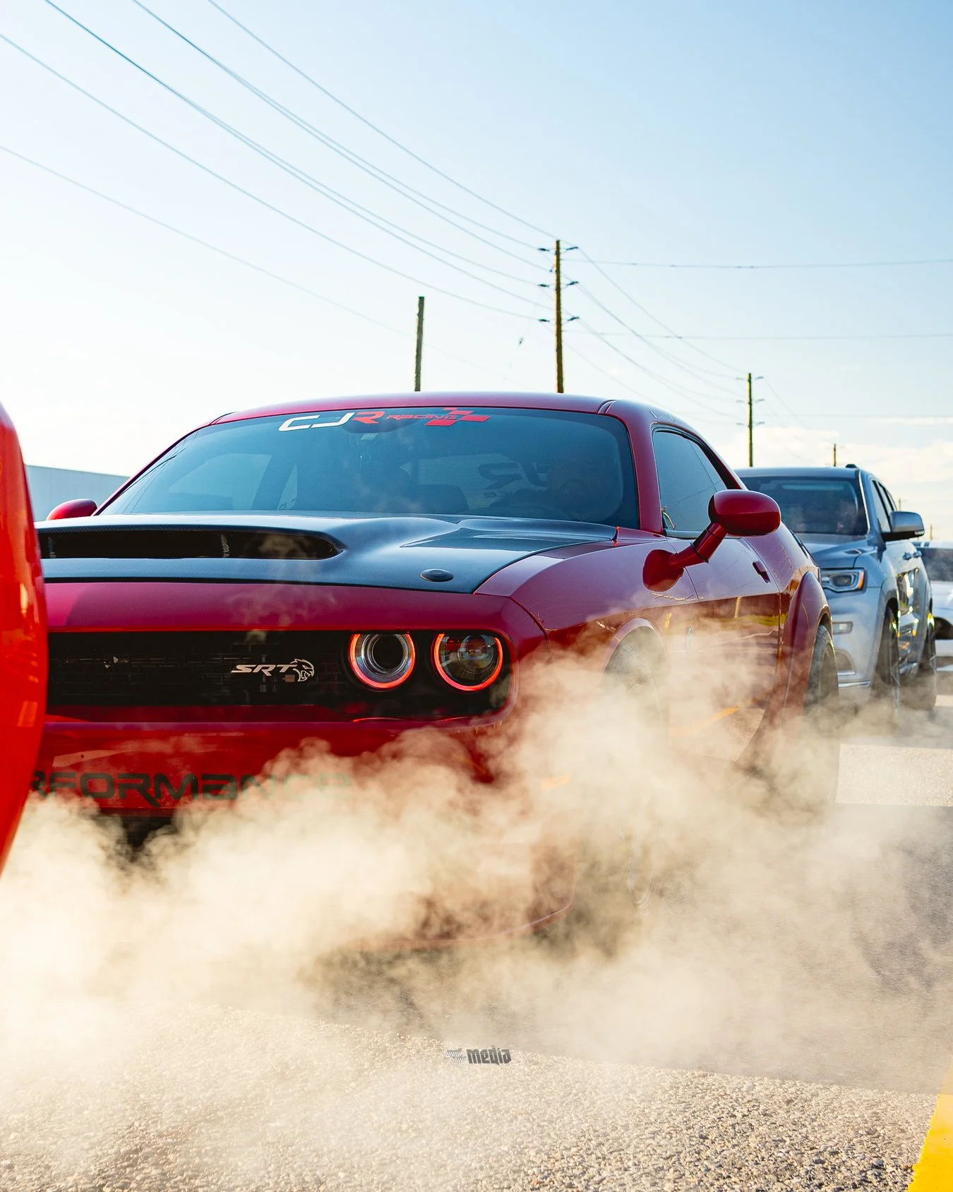 A red Dodge SRT Hellcat car with smoke coming from the tires in a parking lot with other vehicles and utility poles in the background.