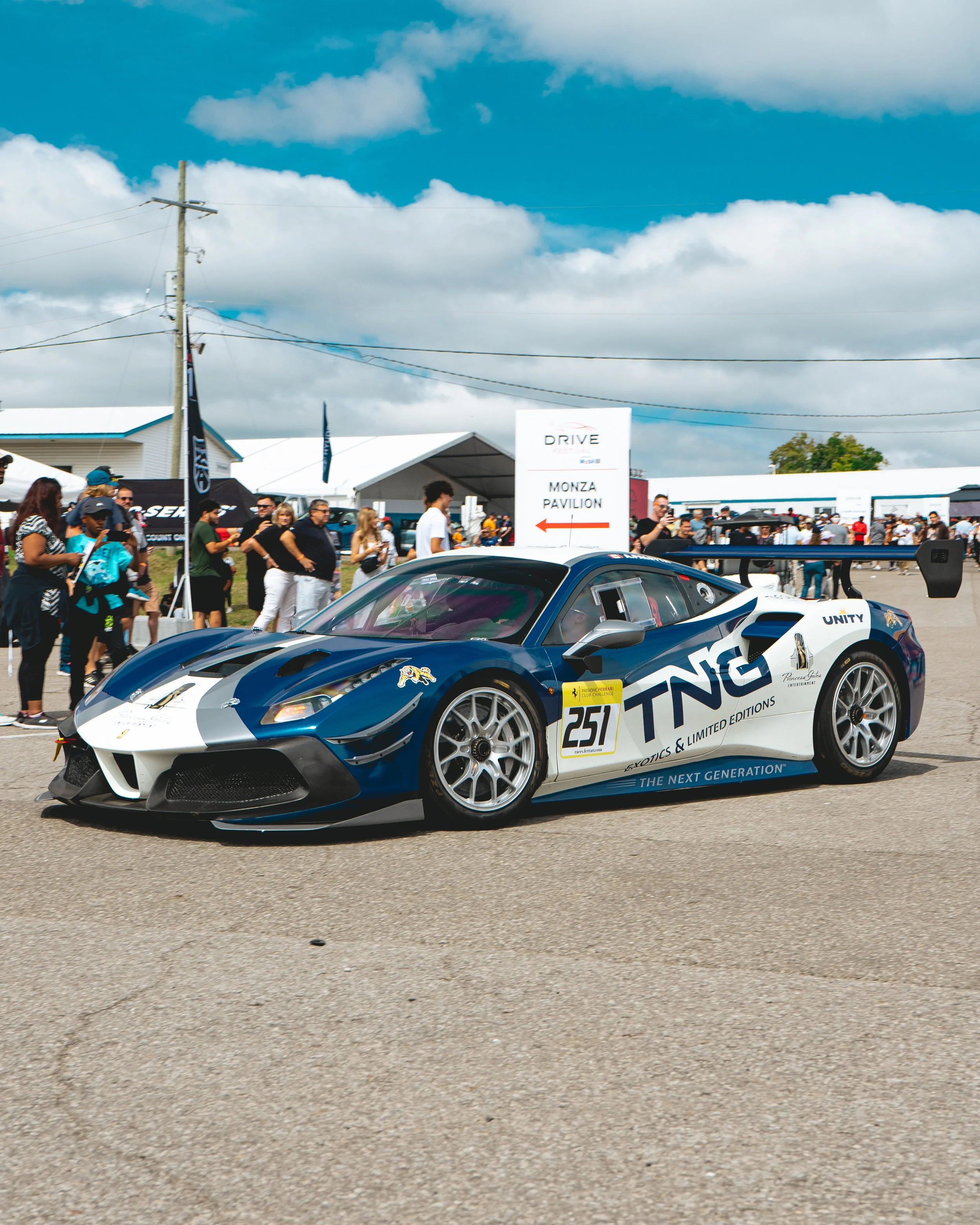 Race car with the number 251 and TNG logo on it parked at a racing event, with a crowd of spectators and tents in the background under a partly cloudy sky.