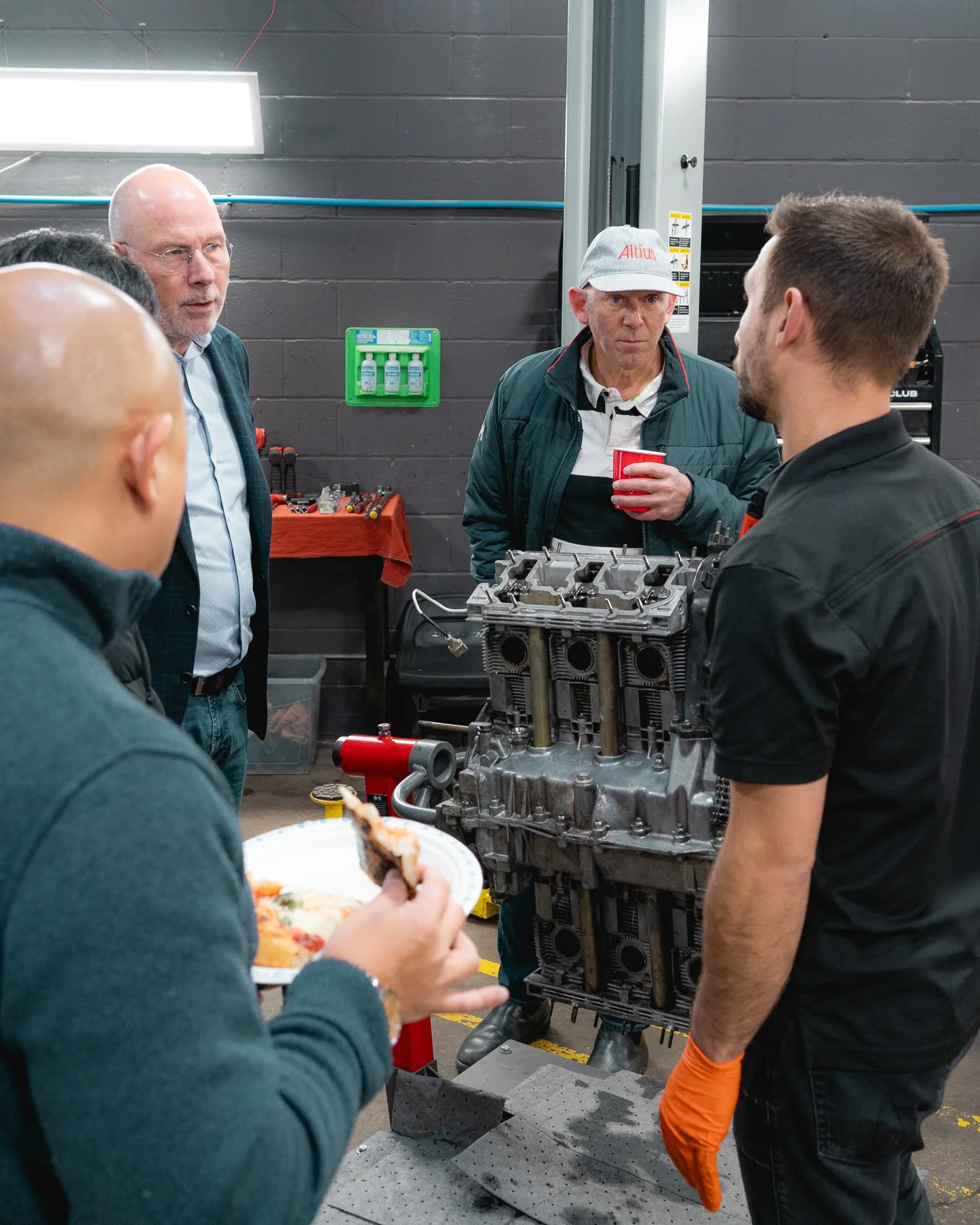 Group of men gathered around an engine block in a garage or workshop, with one man eating pizza and others engaged in conversation.