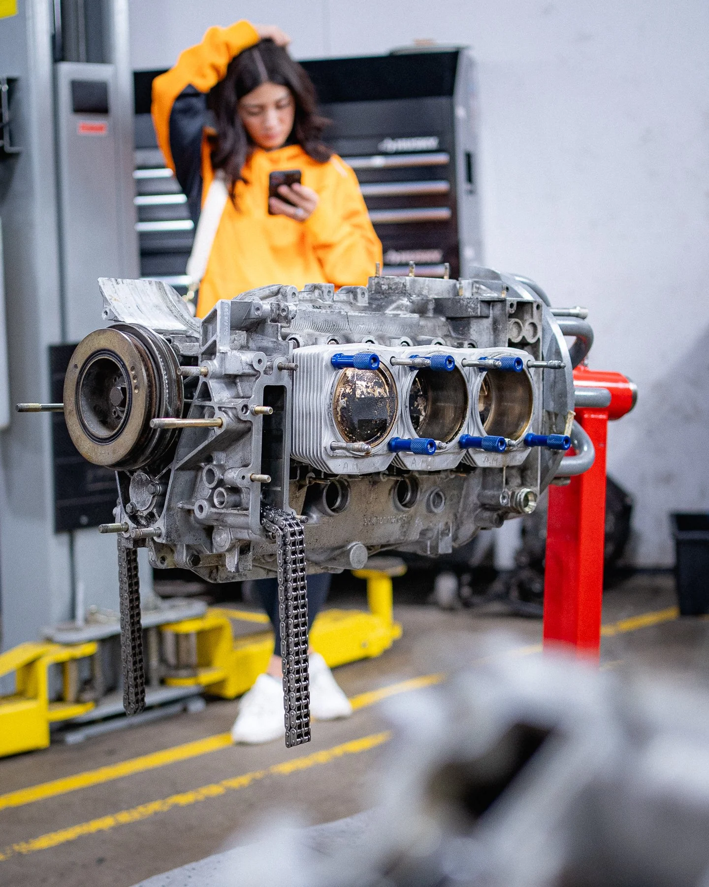 A woman in a yellow and black jacket looks at her phone behind a disassembled car engine mounted on a stand in a garage or workshop.