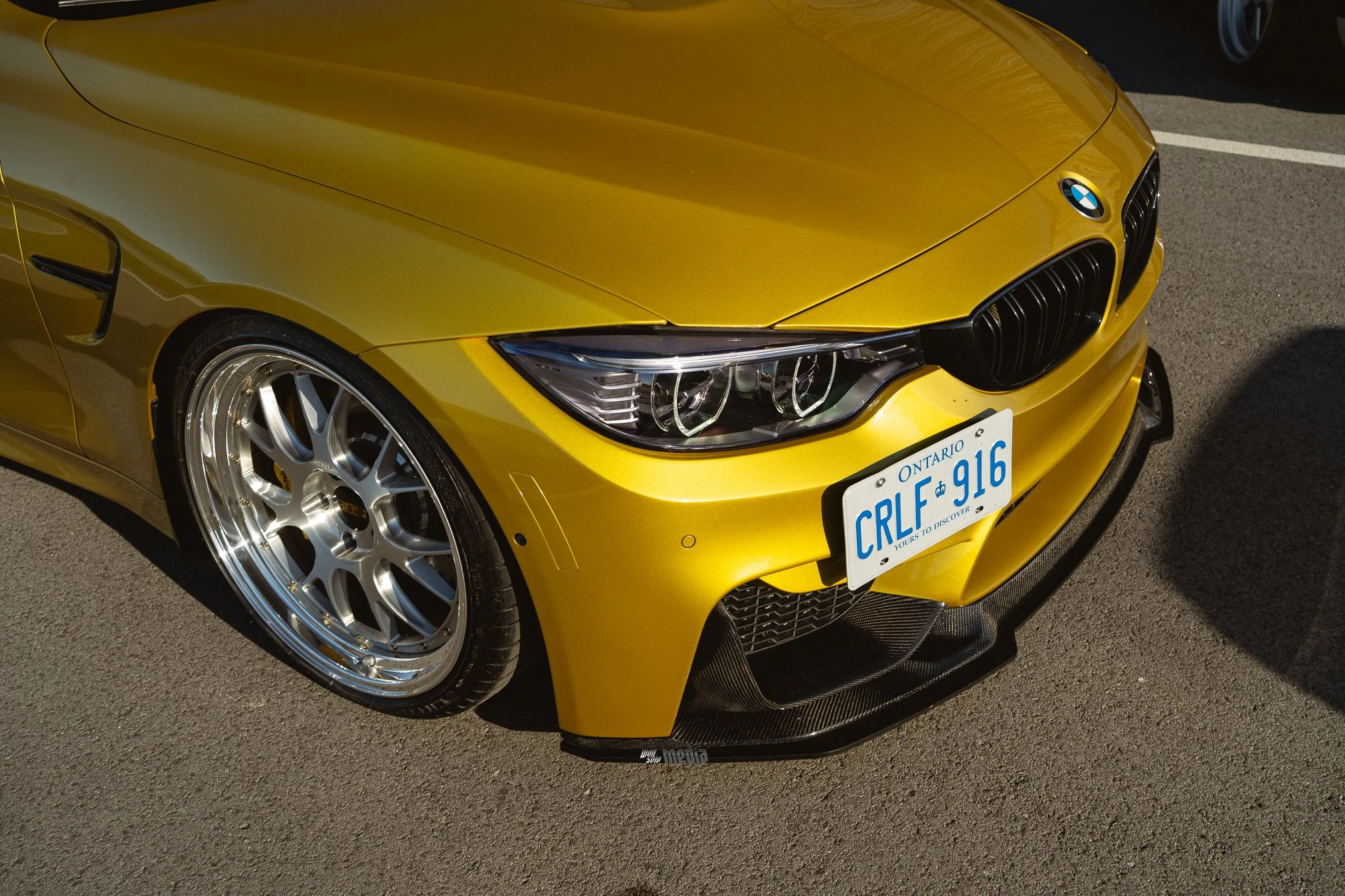 A yellow BMW sports car with chrome wheels parked on a paved surface, showing the front left side including headlight, grille, license plate, and part of the hood.
