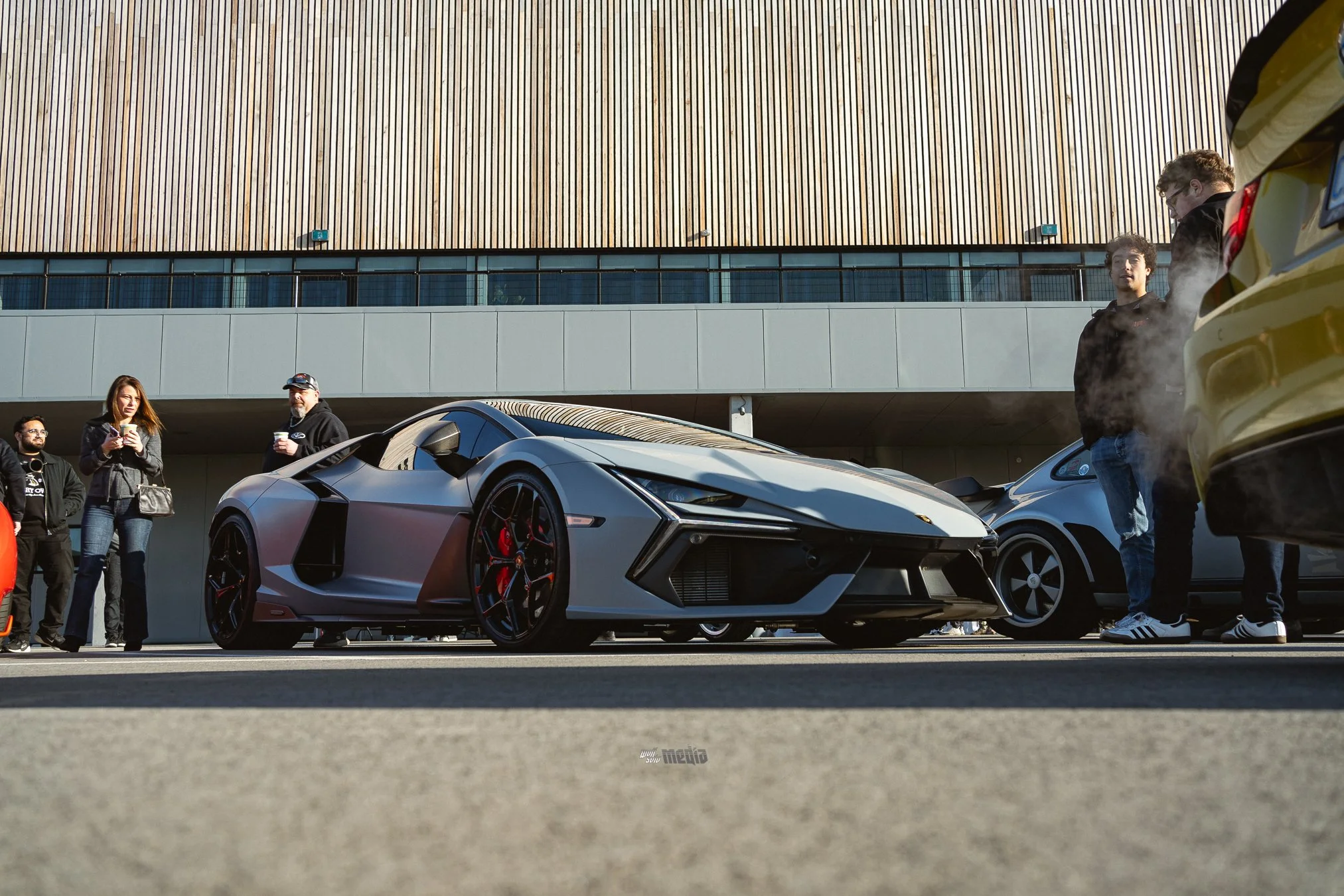A matte gray Lamborghini sports car parked on the street with five people standing nearby, three of whom are watching the car and two are holding drinks, outside a modern building with wooden and glass exterior walls.