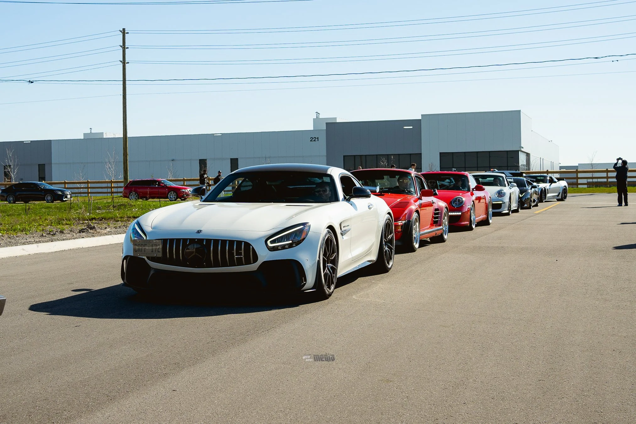 Multiple luxury sports cars parked in a line outdoors with trees, a building, and power lines in the background.