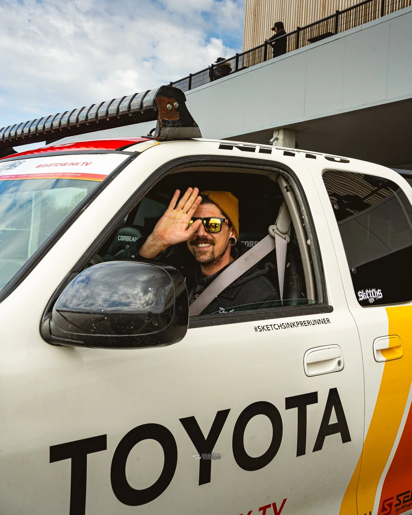 A man wearing sunglasses, a beanie, and a black jacket sitting in a white Toyota race car, waving. The car has a roof scoop and hashtag #SKETCHSINKEPRUNNER on the window, with a crowd and building in the background against a cloudy sky.