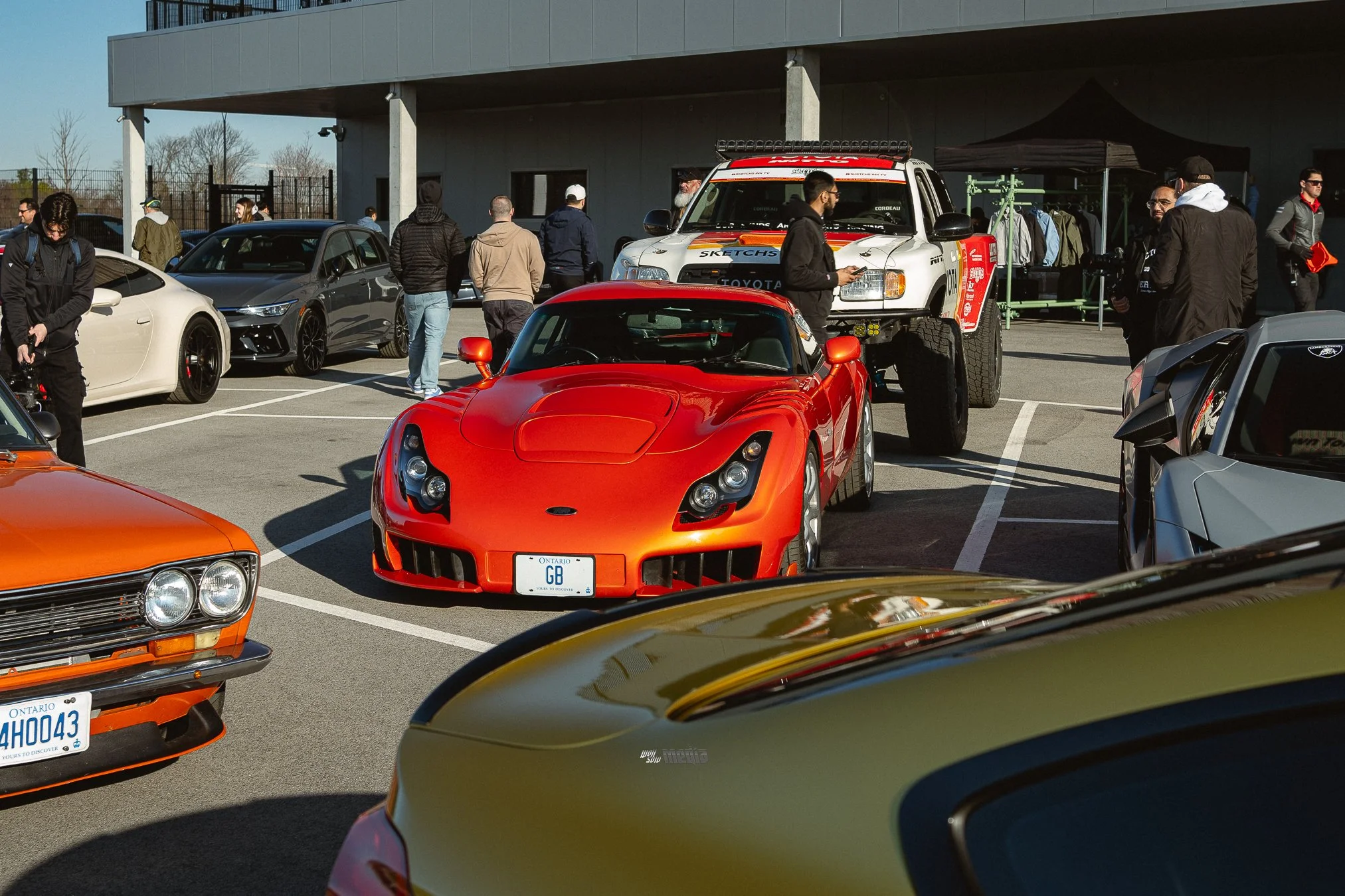 A parking lot filled with various cars, including a prominent red sports car in the foreground, a white rally truck on a trailer, and several other vehicles and people walking around, with a building and some trees in the background.