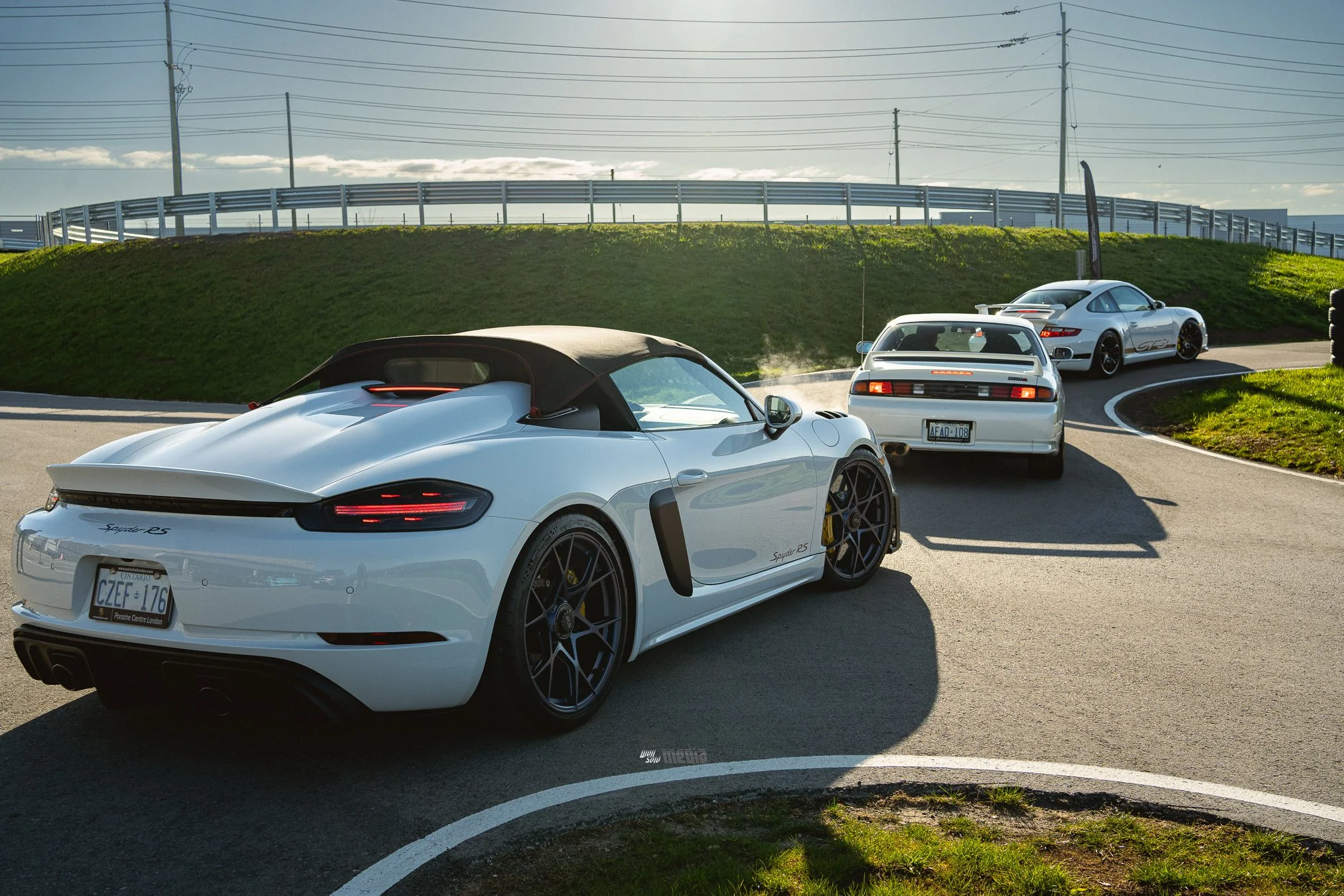 Several white sports cars are parked on a curved parking lot, with a grassy hill and power lines in the background under a partly cloudy sky.