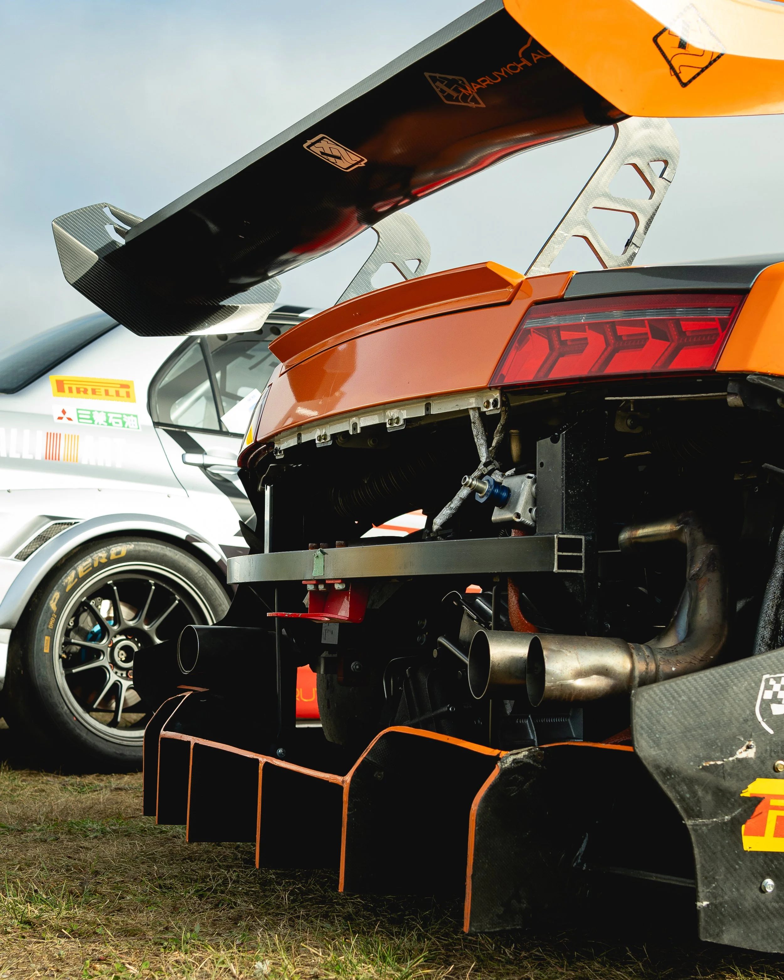 Close-up of a race car's rear end with a large rear wing, exposed exhaust pipes, and aerodynamic elements, parked on grass with another racing vehicle in the background.