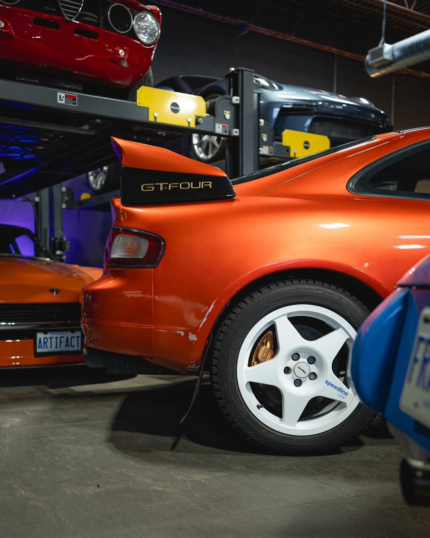 A red Porsche sports car with a black spoiler labeled GT Four, parked indoors with other classic and modern cars around it.