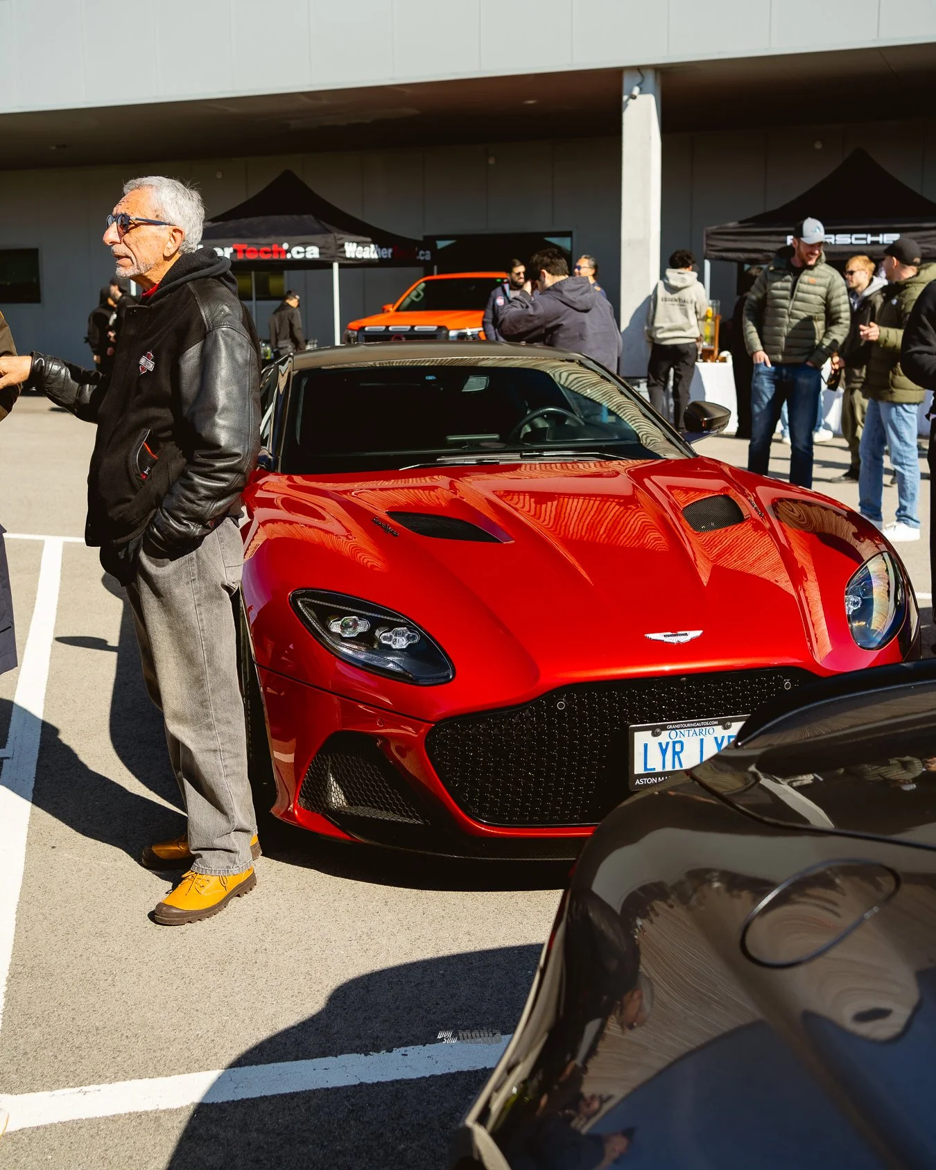 Red sports car with a man in a black leather jacket and gray pants standing next to it during a car show, with several people and tents in the background.