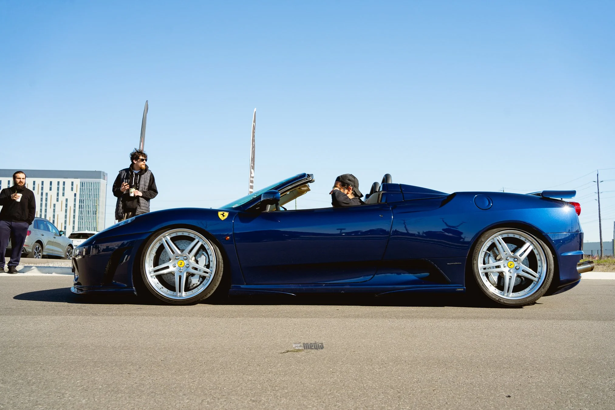 A group of people standing near a shiny blue Ferrari convertible parked outdoors on a clear day. Two individuals are seated in the car, while others are standing nearby, some holding drinks.