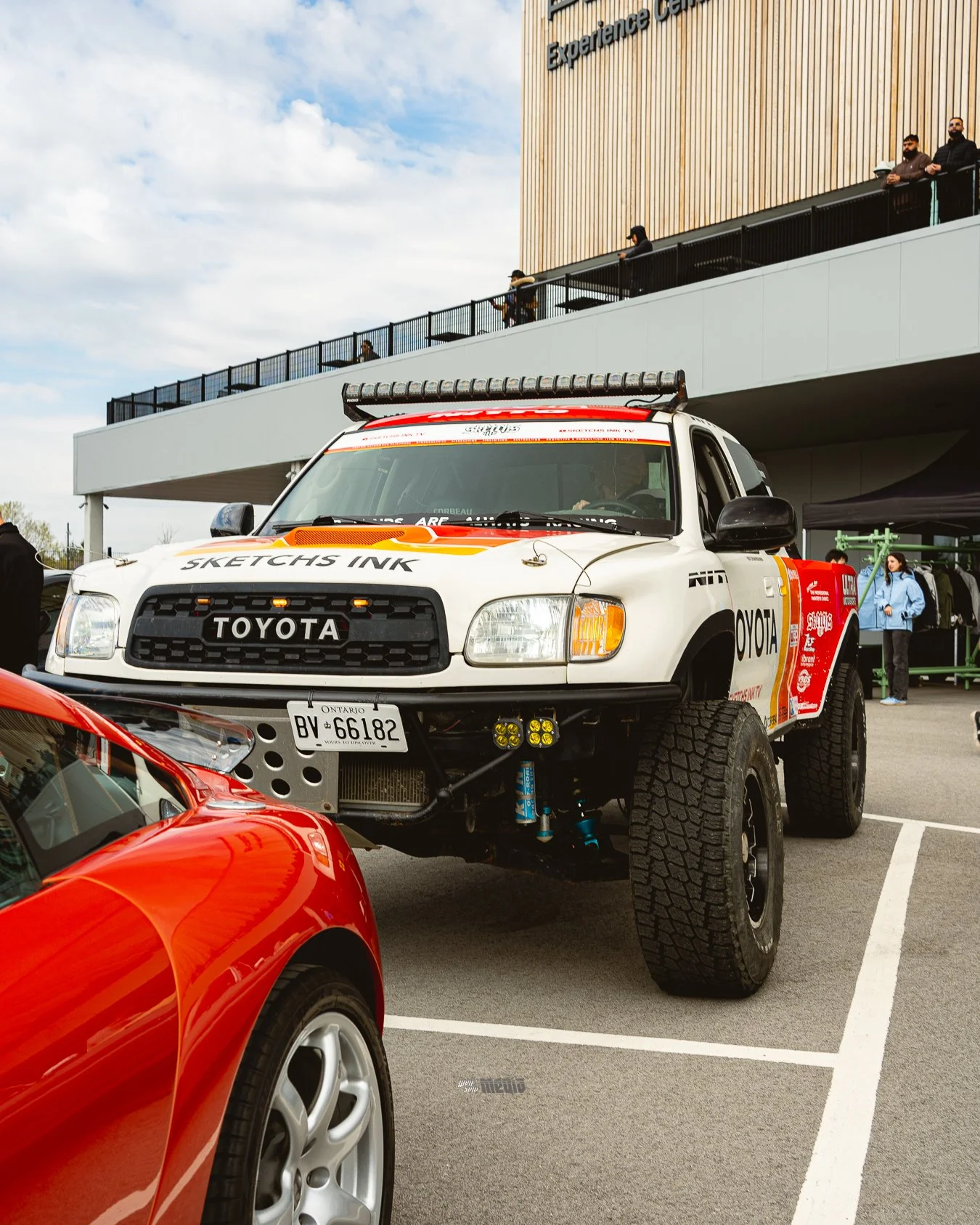 A white Toyota off-road vehicle with large tires and a lifted suspension parked in a lot. In front of it, part of a red sports car is visible. The background shows a building with a wooden facade and a sign that says 'Experience Center'. Some people 